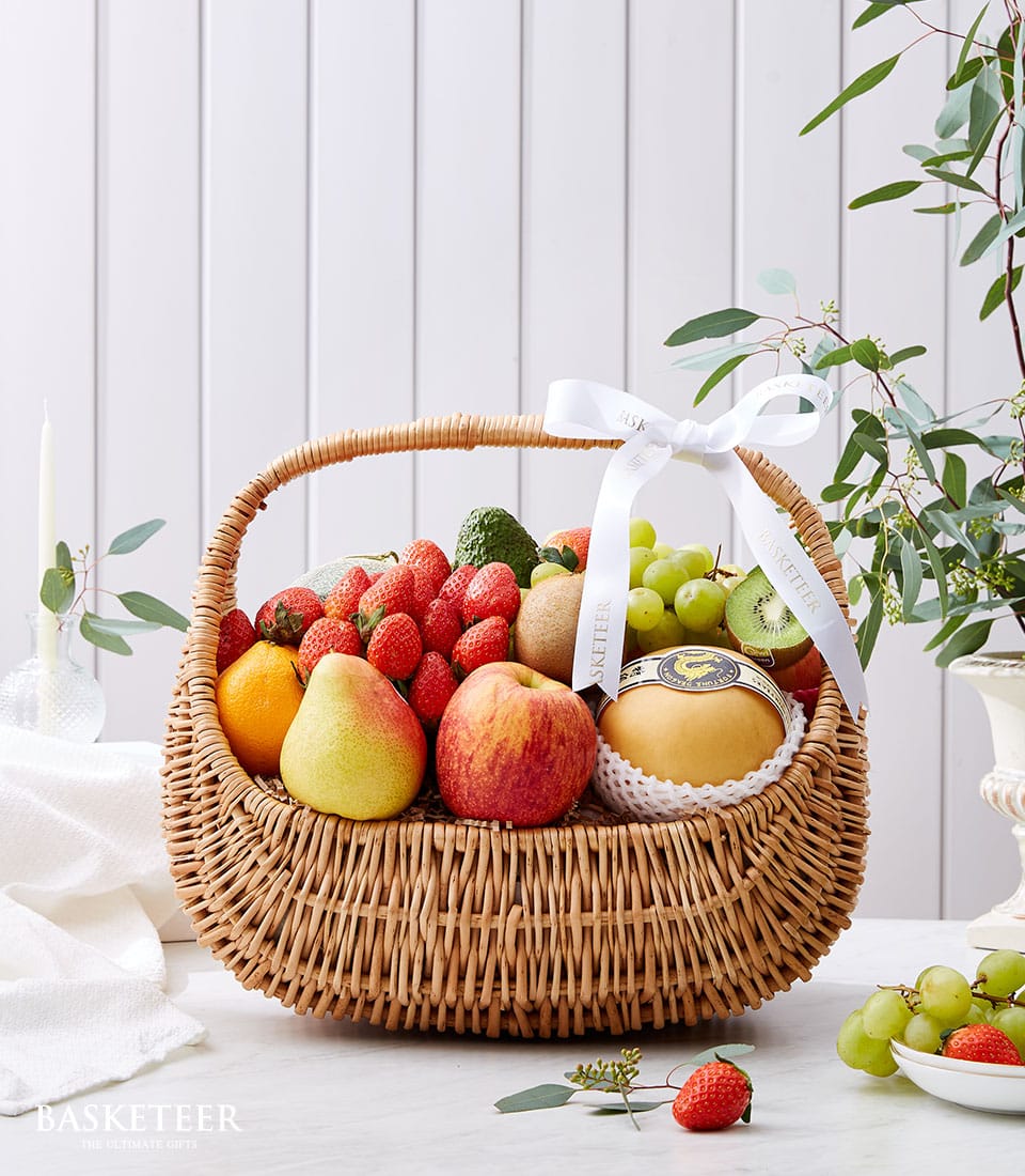 An opulent Basketeer Classic Fresh Fruit Basket overflowing with luscious strawberries, crisp apples, succulent pears, vibrant green grapes, and a perfectly ripened melon. Elegantly adorned with a pristine white ribbon featuring the distinguished Basketeer logo, this exquisite wicker basket is artfully positioned on a pristine white surface. In the background, a softly glowing white candle and an elegant green plant enhance the luxurious ambiance of this inviting scene.