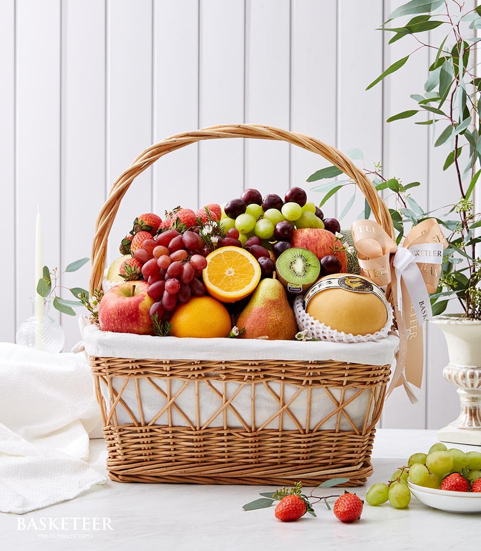 A lavish wicker basket brimming with a selection of Basketeer’s finest premium fruits, including succulent apples, plump oranges, juicy grapes, crisp pears, and exotic kiwi. Nestled among the vibrant array is a gourmet cheese wheel, adding an extra touch of indulgence. The background features a charming white wooden wall adorned with lush greenery, enhancing the scene's natural elegance. A sophisticated ribbon and box proudly displaying the Basketeer logo complete this exquisite presentation—a testament to nature’s luxury at its finest.
