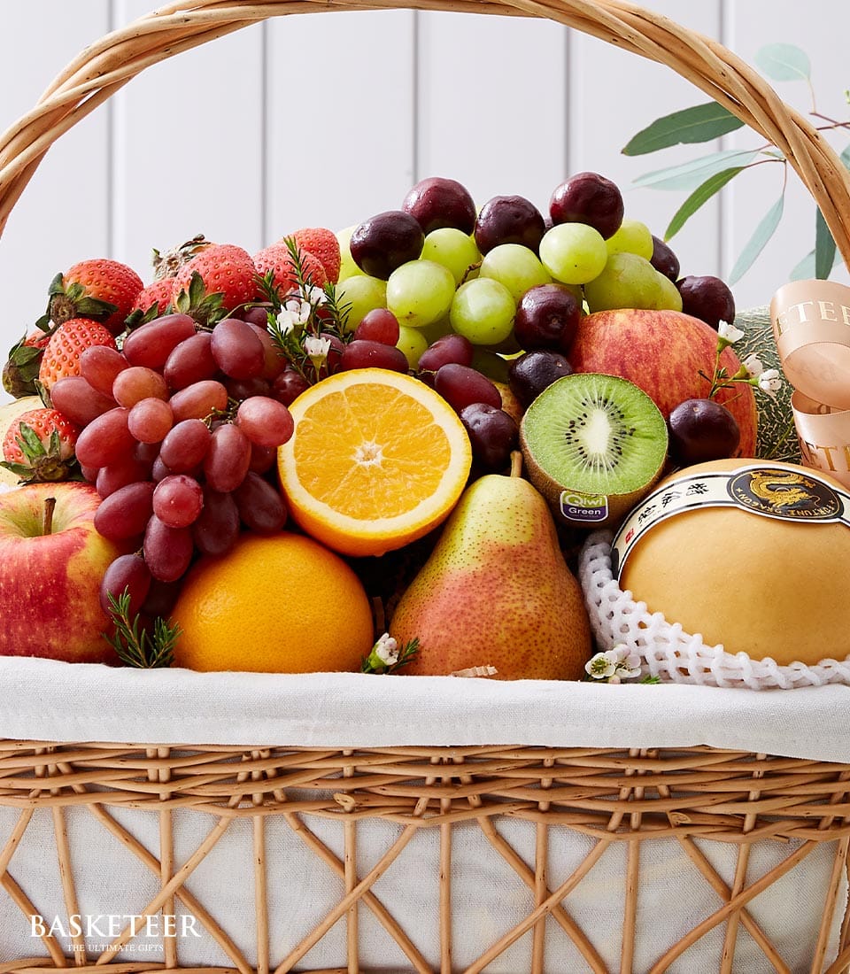A luxurious wicker basket brimming with an exquisite selection of fresh fruits awaits. Inside, you'll find crisp apples, luscious green and red grapes, a sun-kissed orange, succulent strawberries, a vibrant kiwi, and a delectable pear. Nestled amongst these finest fruits is a round of elegantly wrapped cheese. This opulent arrangement forms one of Basketeer's Premium Fruit Gift masterpieces. The scene is enshrined by verdant green leaves in the background, enhancing the natural elegance and inviting presentation inherent in the Basketeer brand experience.
