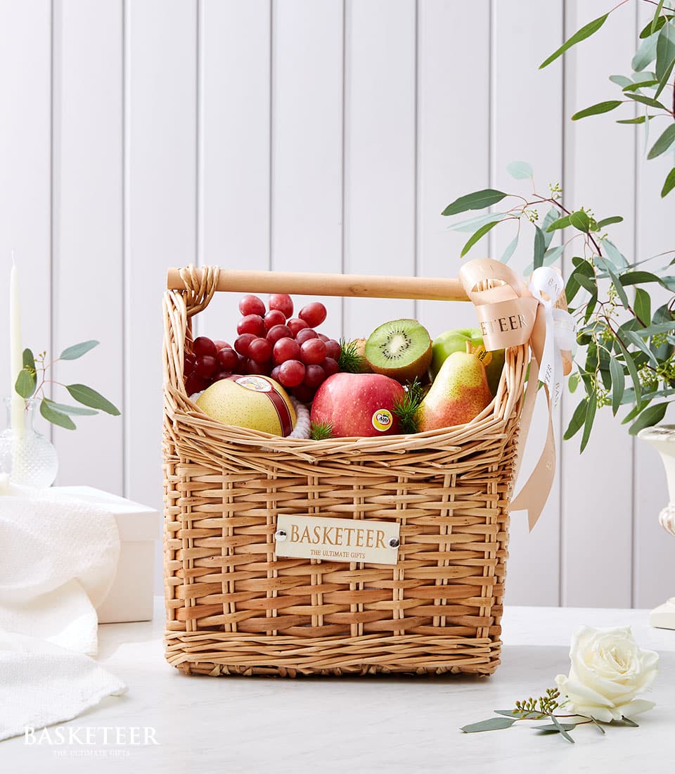 An exquisite wicker basket brimming with an opulent selection of fresh fruits — succulent red grapes, vibrant kiwi, crisp red and green apples, and a perfectly ripe pear — rests elegantly on a pristine white surface. The lavish offering is adorned with a sophisticated ribbon and proudly bears the distinguished 