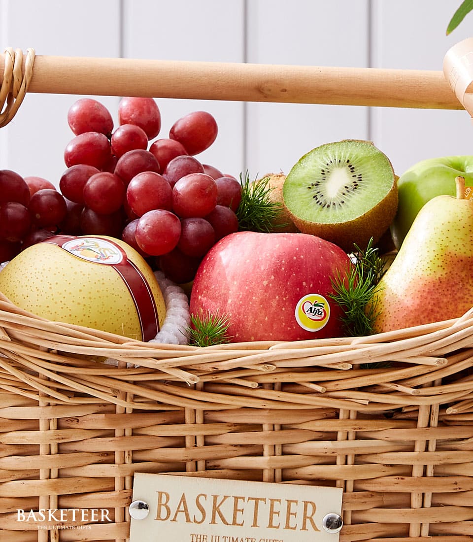 A luxurious wicker basket, elegantly designed with a light wooden handle, displays an opulent assortment of fresh, vibrant fruits. Nestled within are luscious red grapes, a succulent half-cut kiwi, a crisp green apple, a juicy red apple, and an exotic Asian pear. The basket features a bespoke label on the front proudly displaying the distinguished 