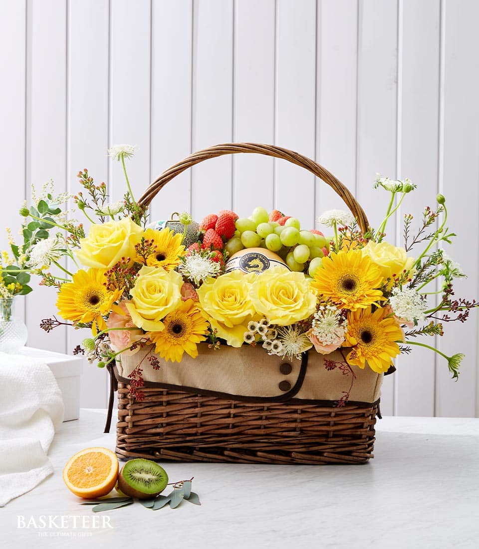 An opulent Sunny Blooms Fruit Basket by Basketeer, elegantly adorned with a vibrant bouquet of yellow roses, sunflowers, and lush greenery. This luxurious arrangement is complemented by an array of fresh fruits such as plump grapes, succulent strawberries, refreshing kiwi slices, and zesty orange wedges. The wicker basket boasts the distinguished Basketeer logo on a ribbon and rests gracefully on a pristine white table against a backdrop of light wooden paneling, creating an inviting and serene ambiance.
