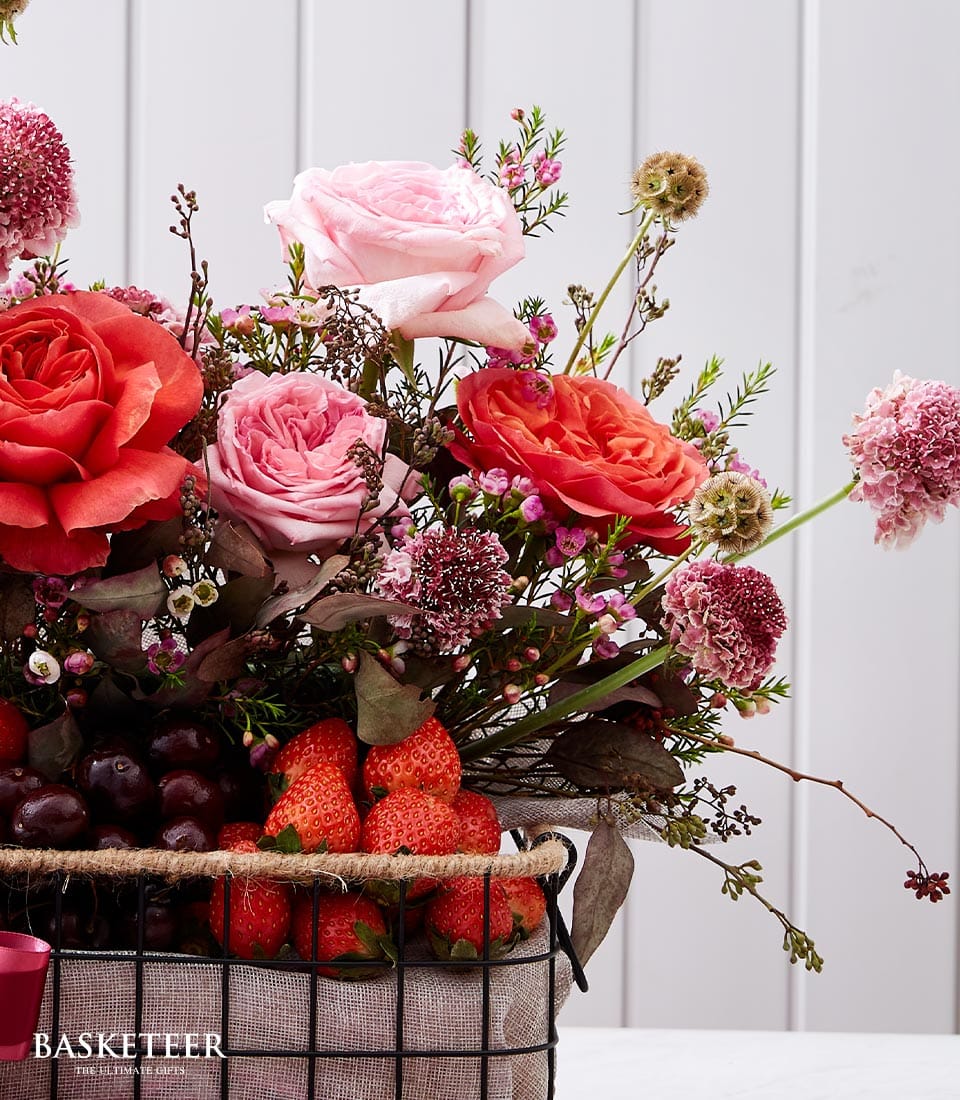 The Basketeer Sweetheart Fruit Floral Basket is an exquisite fusion of vibrant red, pink, and purple blossoms paired with the succulent freshness of strawberries and grapes. The carefully arranged flowers emanate luxury, nestled within a basket adorned with a tasteful Basketeer ribbon. Against a sophisticated light backdrop accentuated by subtle vertical lines, this elegant composition invites admiration and indulgence.
