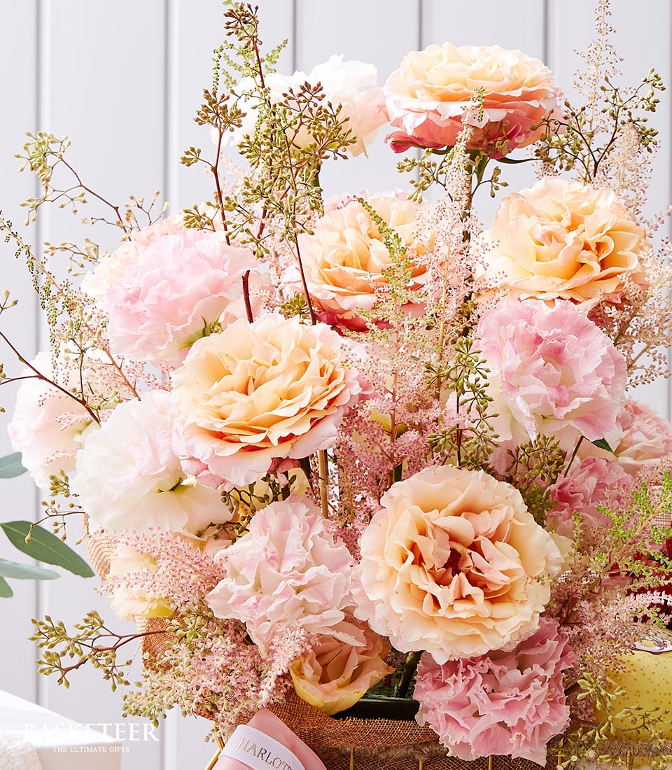 A luxurious, inviting bouquet of soft pastel flowers, featuring exquisite peach and blush roses, delicate pink hydrangeas, and airy green foliage. These stunning blossoms are artfully arranged in a woven fruit basket adorned with a tasteful Basketeer ribbon. The scene is set against an elegant light-colored vertical paneled wall, enhancing the sophistication of the arrangement.