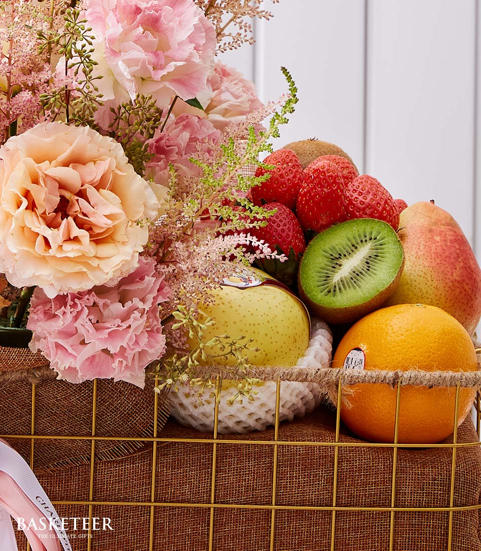 A lavish basket, bedecked with a symphony of assorted fruits—succulent strawberries, a vibrant kiwi, a luscious pear, a zesty orange, and a crisp apple—nestles amidst an opulent array of pink and peach flowers. Carefully curated within an elegant brown mesh basket framed by gleaming yellow metal that proudly spells 