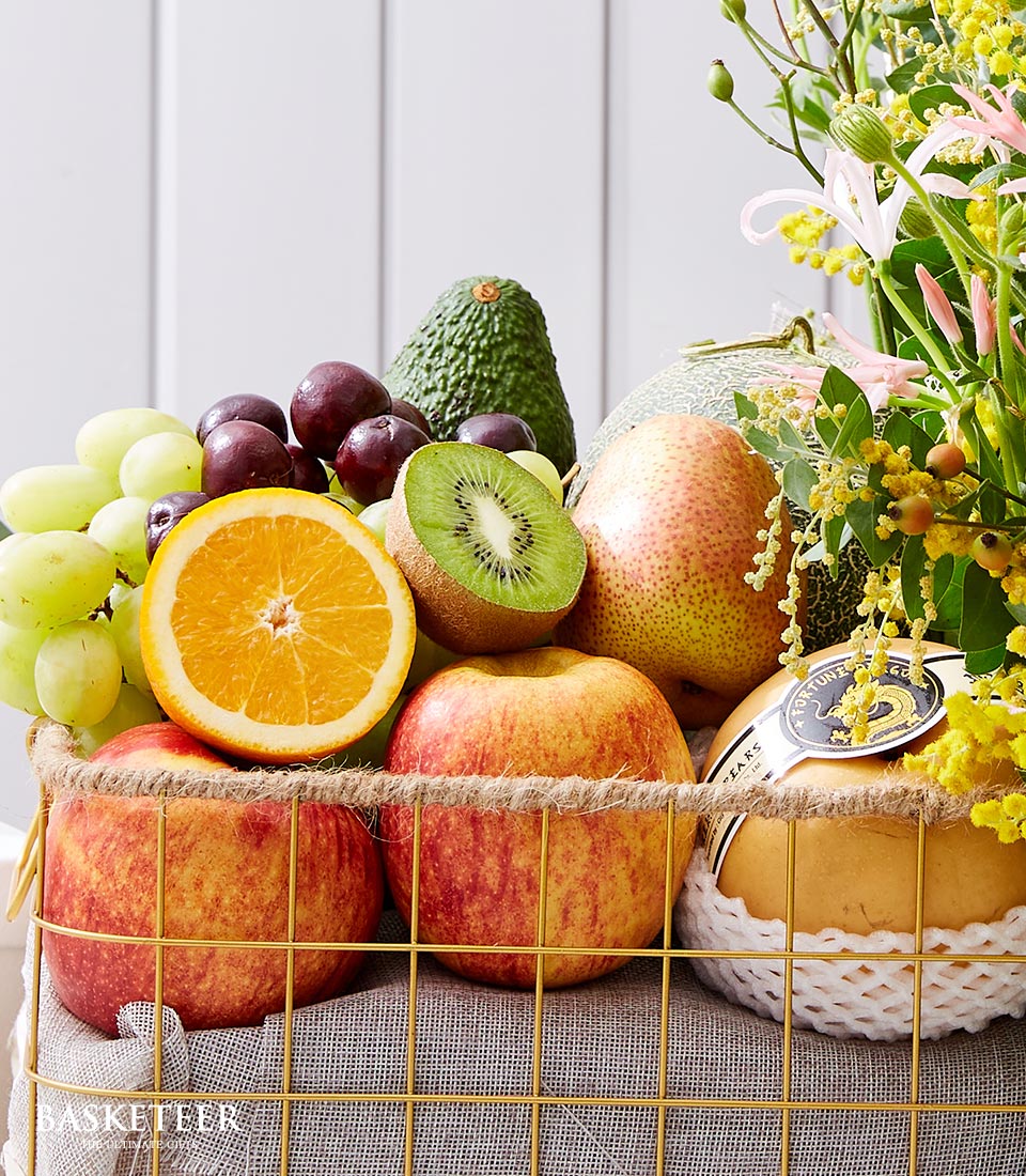 A luxurious Basketeer fruit flower basket brims with an exquisite assortment of the finest fresh fruits, including crisp apples, succulent kiwi, plump grapes, a perfectly ripe avocado, a zesty orange half, and a luscious pear. Nestled among the vibrant bounty is a wrapped golden cheese wheel and an elegant bouquet of flowers that brings an additional splash of greenery and color to this opulent display. The scene exudes warmth and sophistication, inviting you to indulge in Basketeer's signature blend of premium produce and artisanal flair.