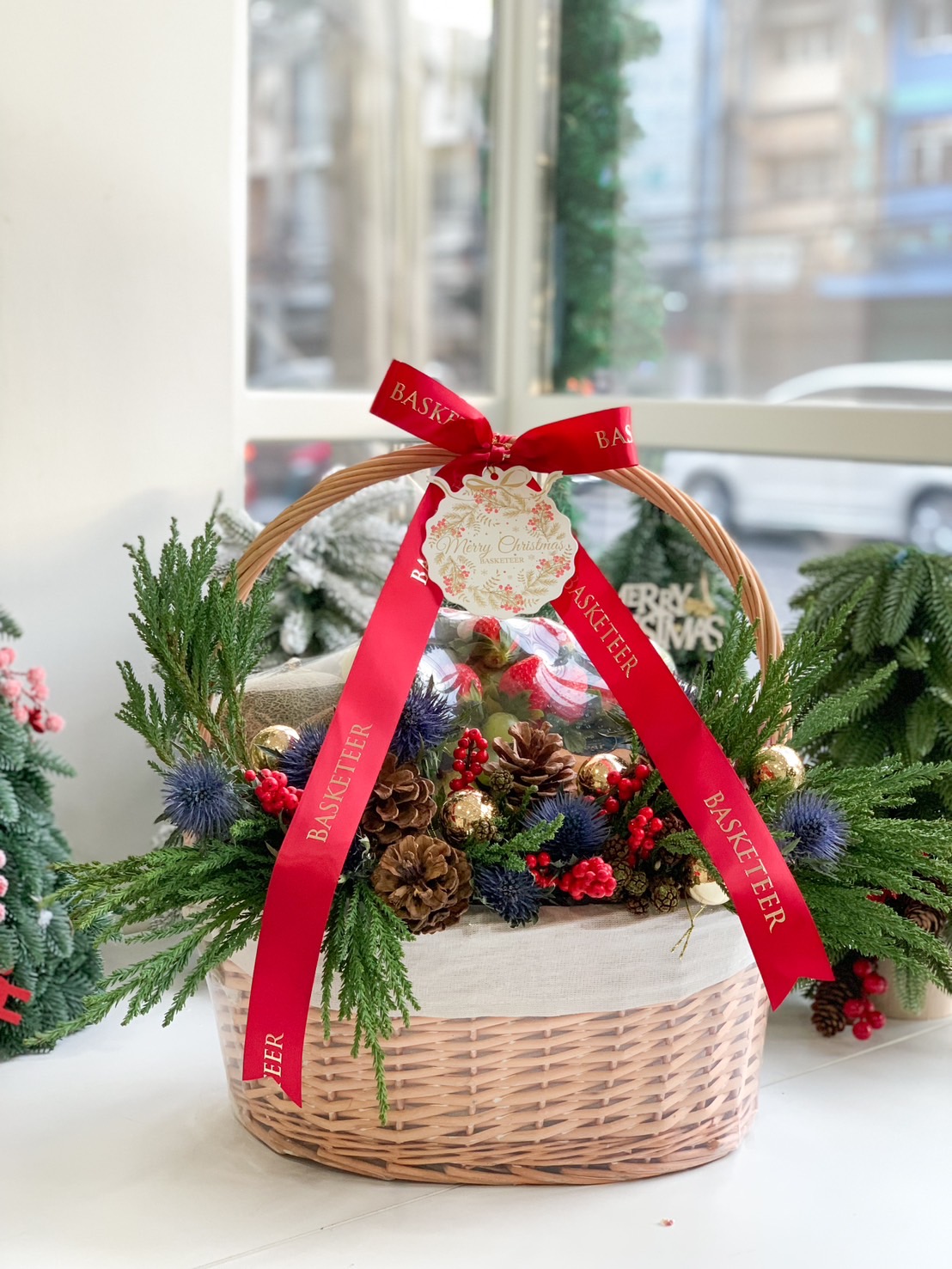 Deluxe holiday fruit basket with fresh apples, avocados, grapes, strawberries, and festive pinecone accents.