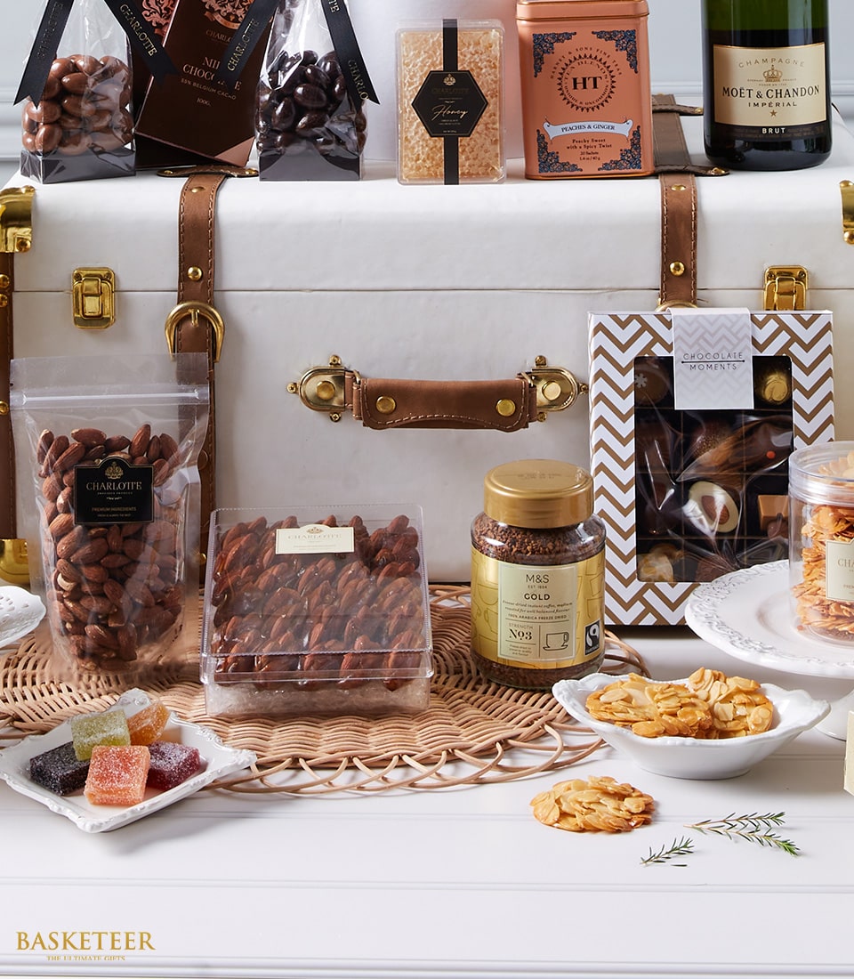 A luxurious Basketeer gift hamper featuring an exquisite selection of assorted chocolates, premium nuts, artisanal honey, gourmet popcorn, fruity jellies, and a prestigious bottle of Moët & Chandon champagne. These indulgent treats are meticulously arranged within and around a sophisticated white leather trunk adorned with tan straps. The elegant presentation reflects Wine Connoisseur's Gourmet Elegance. A delicate ribbon bearing the Basketeer logo cascades over the trunk, adding to the opulent allure of this enchanting offering.