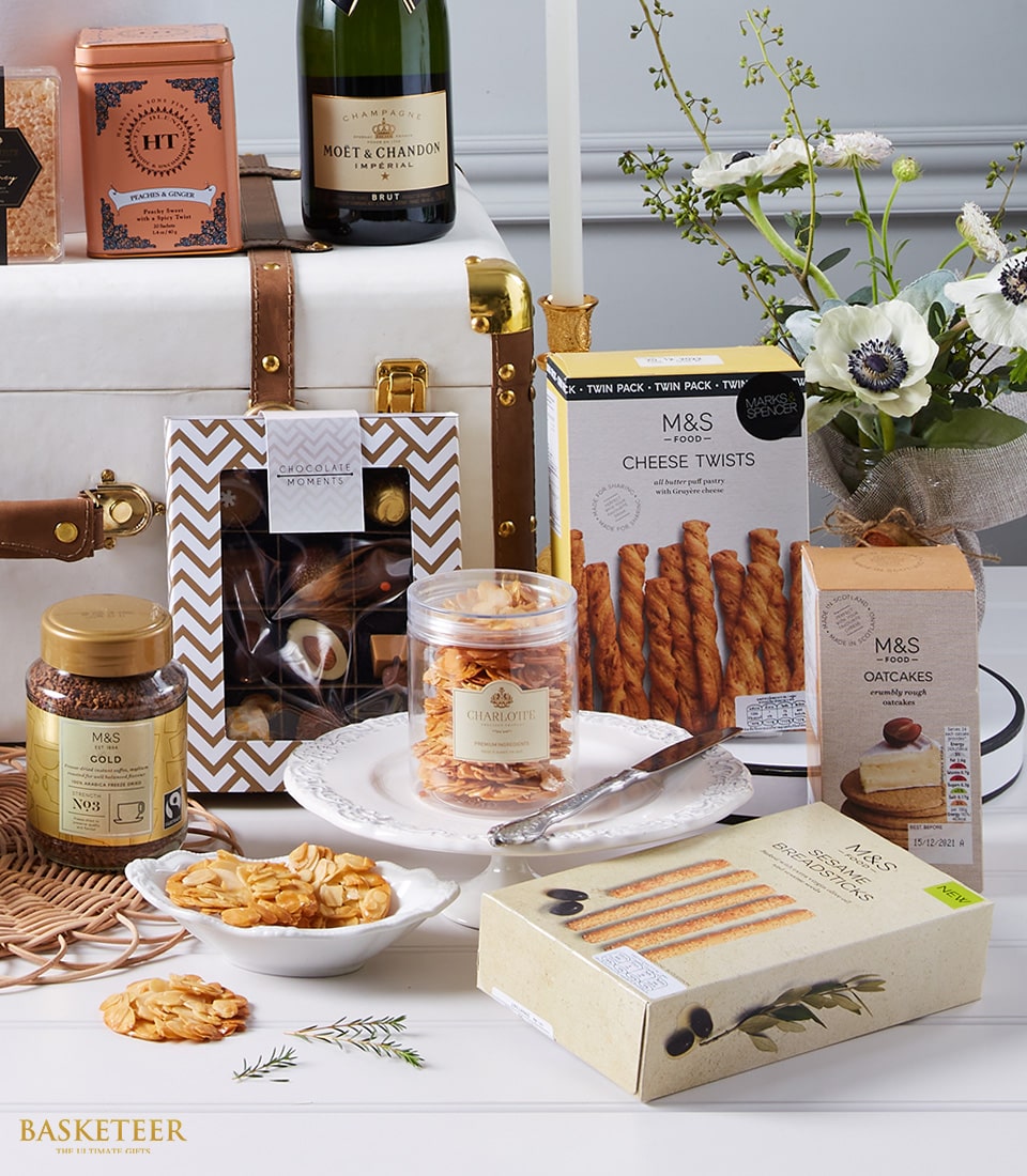 An opulent gift arrangement epitomizing gourmet elegance from Basketeer features a bottle of Moët & Chandon champagne, accompanied by an exquisite selection of M&S delicacies including cheese twists, oatcakes, chocolate biscuits, and honey. These indulgent treats are artfully presented on a pristine white table adorned with fresh flowers. In the background, a luxurious white hamper trimmed in fine leather and featuring the distinguished Basketeer logo completes this inviting scene.