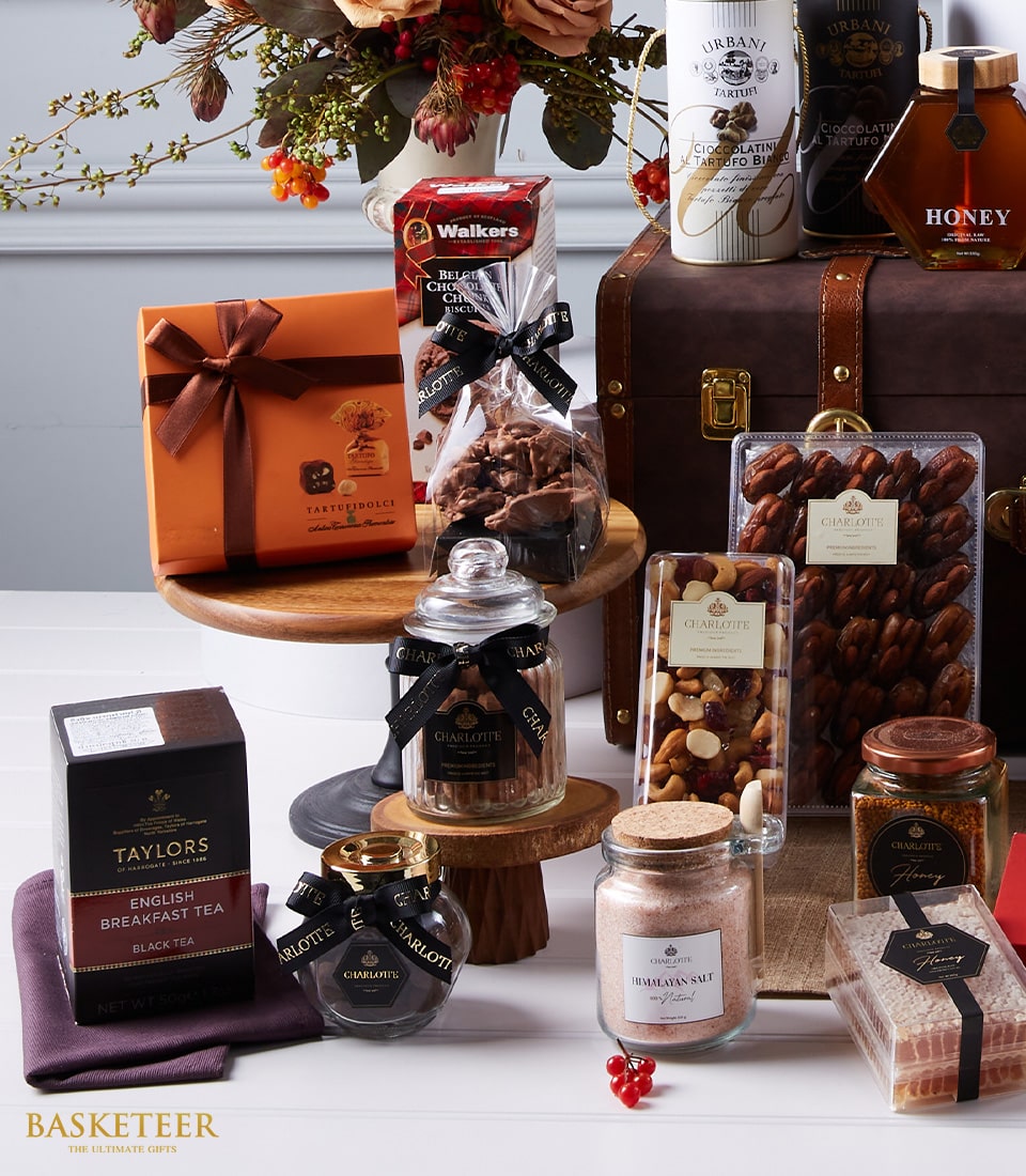 An opulent Basketeer Premium Wine and Tempting Treats Hamper, exquisitely curated for the discerning connoisseur. This luxurious selection features an array of gourmet delights: assorted artisanal chocolates, aromatic English breakfast tea, rich honey, delectable Turkish delight, a medley of premium mixed nuts, pristine Himalayan salt, indulgent Walkers milk chocolate toffees, and decadent Champagne truffles. These treasures are thoughtfully displayed on refined wooden stands and nestled within an elegant trunk adorned with the distinguished Basketeer logo. A lush floral arrangement graces the background, elevating the scene into one of pure sophistication and inviting grandeur.