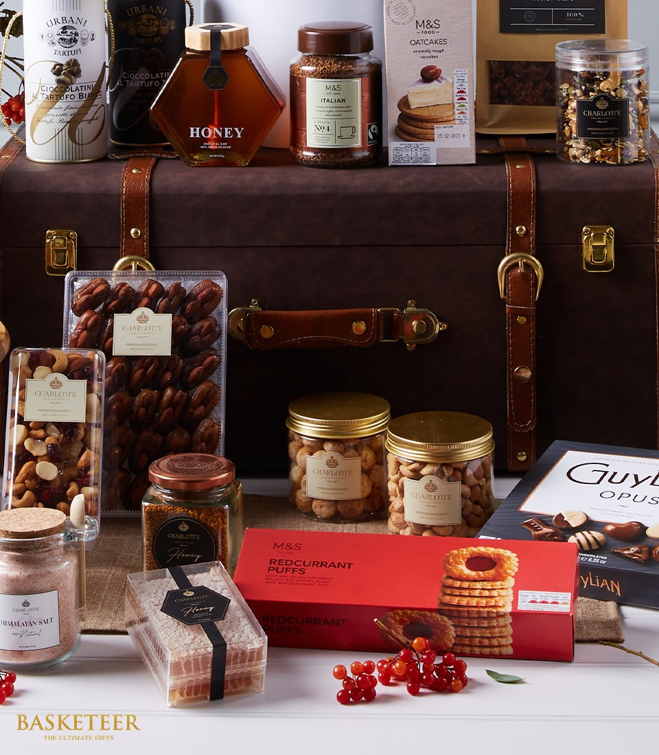 A luxurious Basketeer Premium Wine and Tempting Treats Hamper, elegantly presented in a vintage-style leather trunk adorned with the Basketeer logo. This exquisite hamper features a curated selection of gourmet delights including artisanal honey, rich chocolate, premium nuts, delectable oatcakes, mouthwatering currant puffs, and sumptuous jam. The scene is accentuated with small red berries scattered around for an added festive touch, creating an inviting and opulent atmosphere perfect for any celebration.