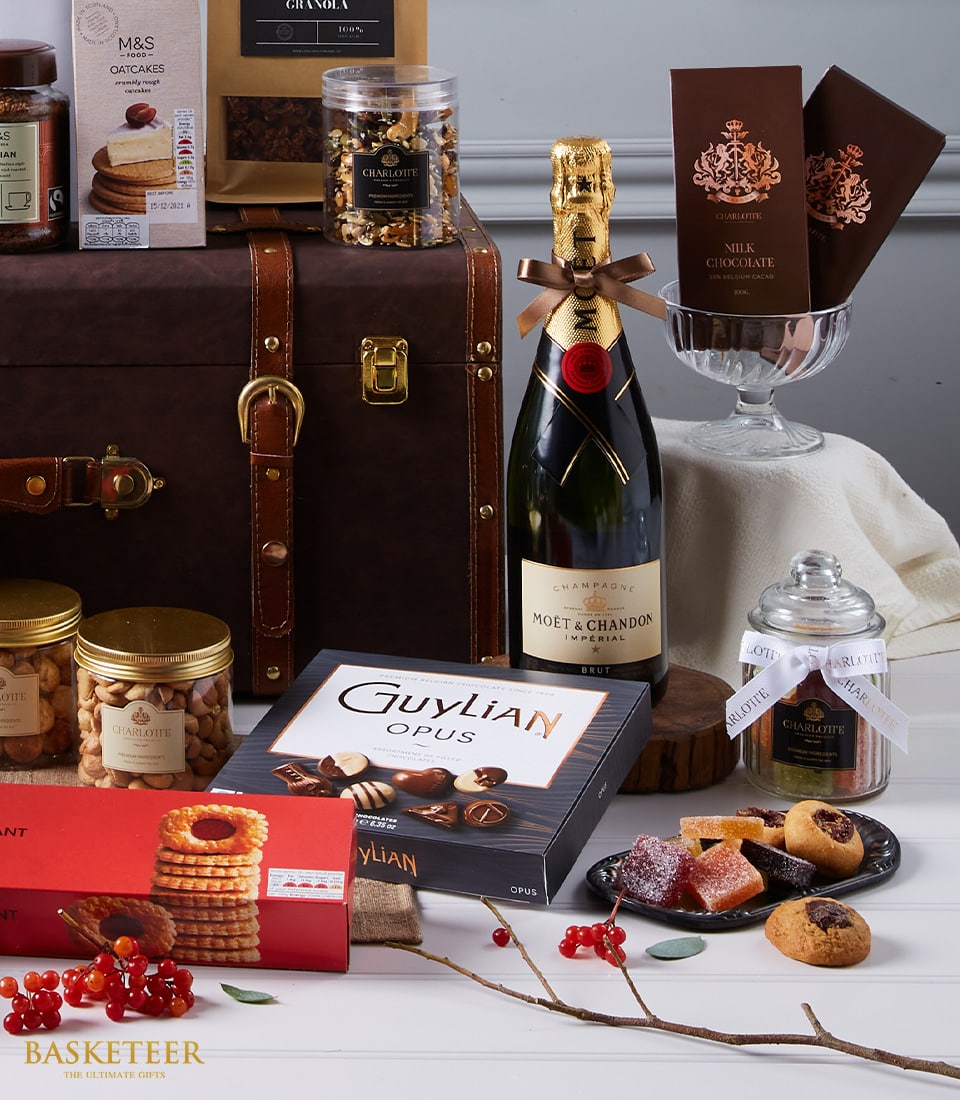 An opulent gift set showcasing a bottle of Moët & Chandon champagne, an assortment of decadent chocolates, gourmet cookies, fine nuts, and exquisitely preserved fruits. Each item is meticulously arranged in a sophisticated leather trunk with elegant glass jars, all presented on a pristine white surface. This lavish Premium Wine and Tempting Treats Hamper by Basketeer is the epitome of luxury for any celebration. The 