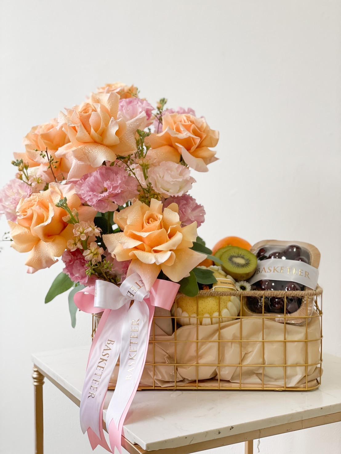 A decorative basket filled with a bouquet of orange roses and pink flowers on one side. The other side features an assortment of foods, including kiwi, grapes, and a jar labeled 