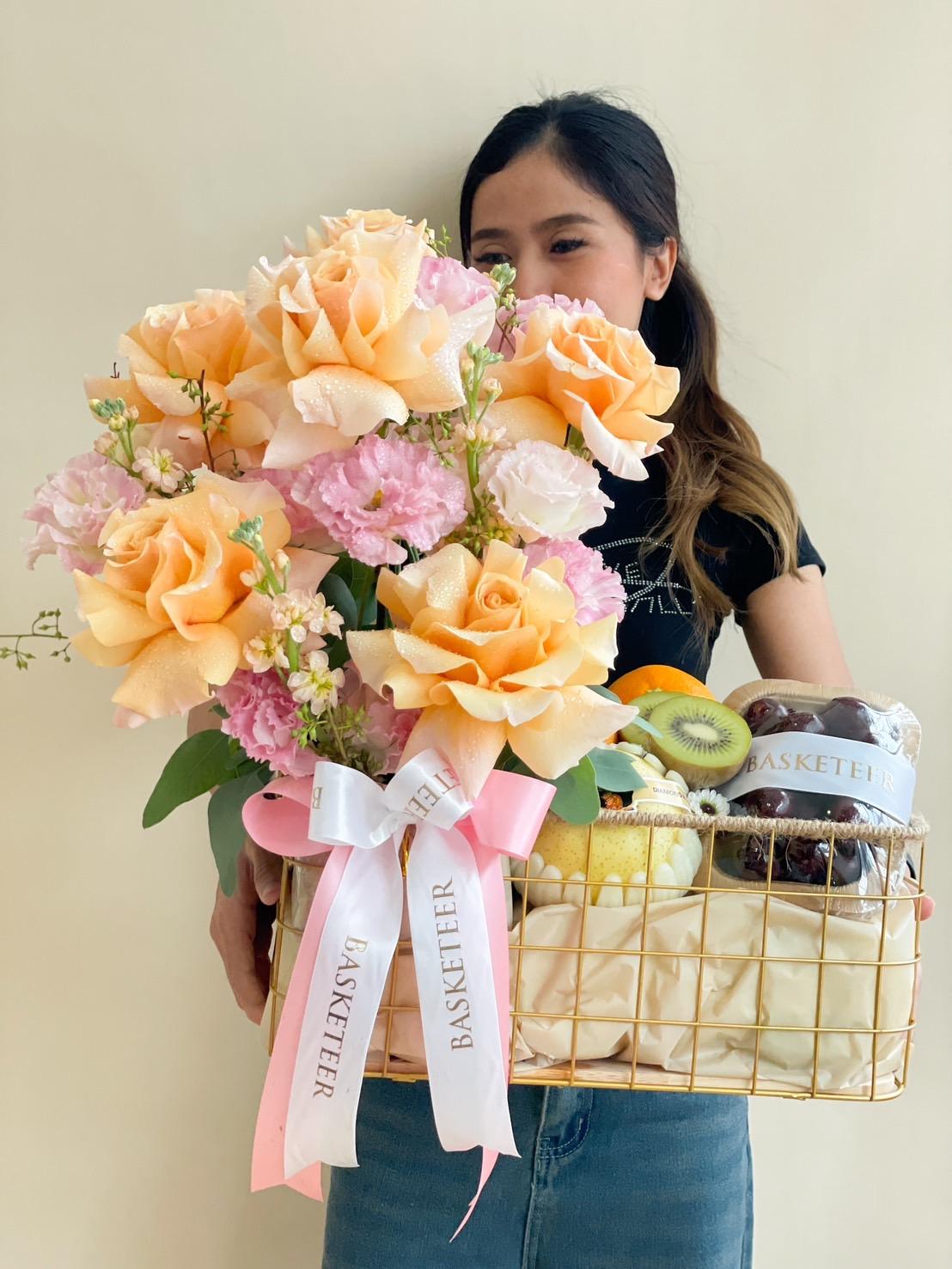 A woman holding a gold wire basket filled with large peach roses, pink flowers, and various fruits including kiwis and apples. The basket, adorned with a white ribbon reading 
