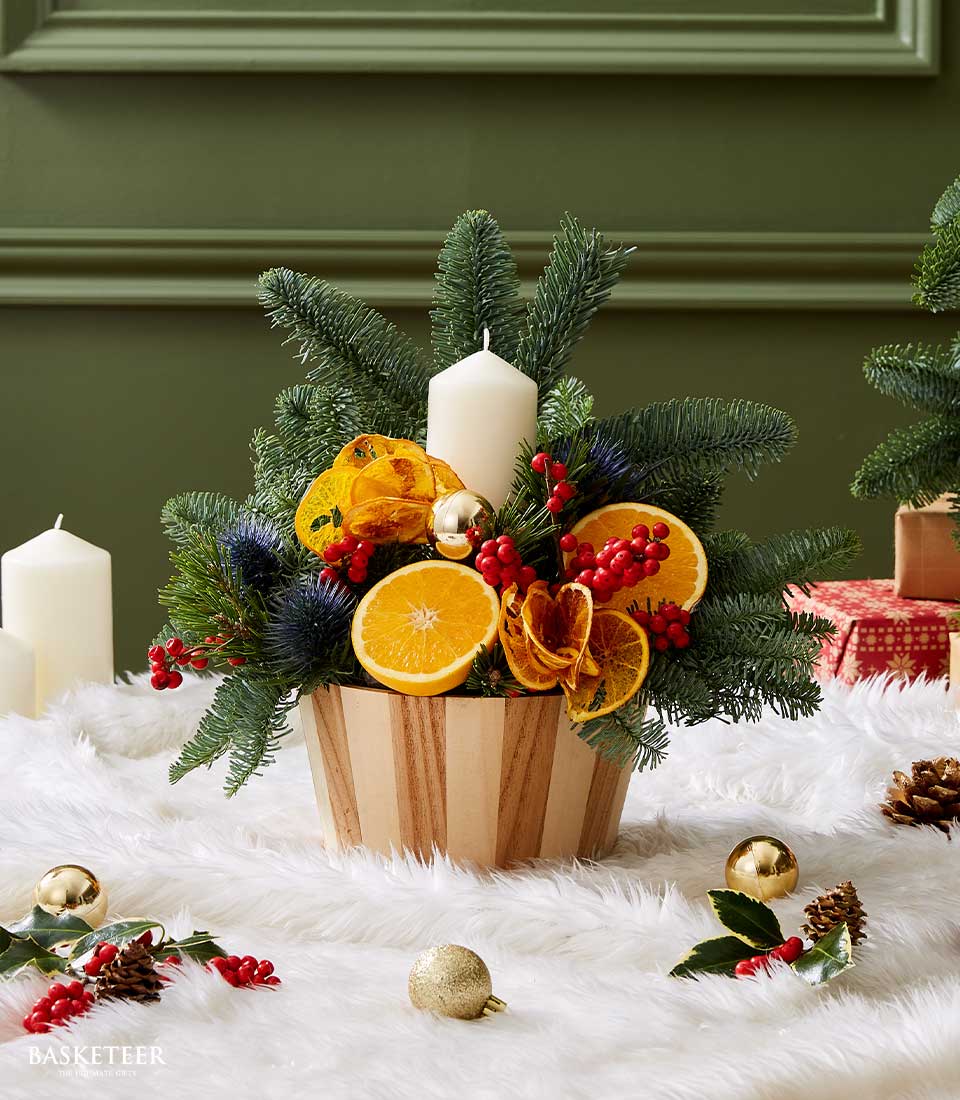 Christmas centerpiece with a white candle, dried orange slices, red berries, and festive greenery in a wooden base.