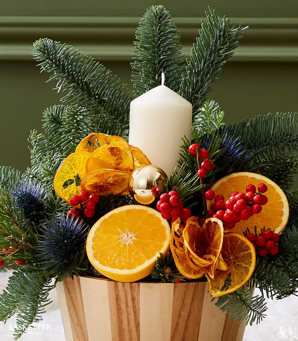 Close-up of Christmas centerpiece with a white candle, dried orange slices, red berries, and festive greenery in a wooden base.