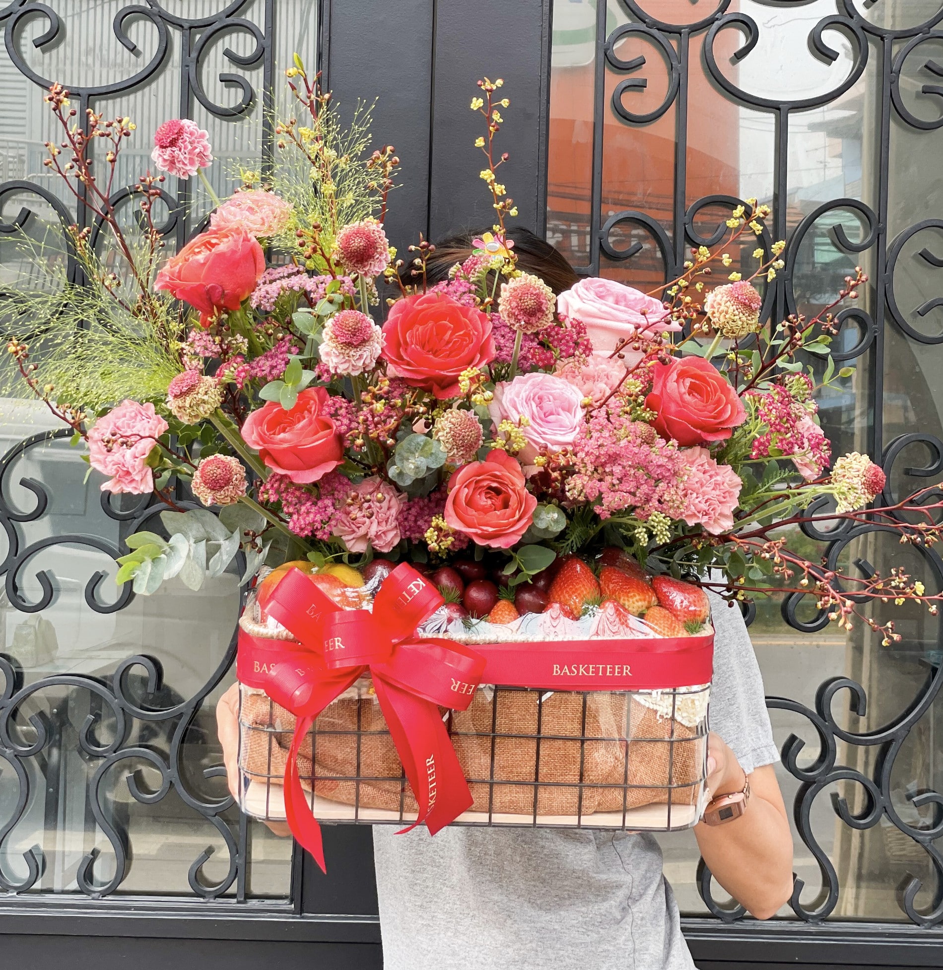 A person holding a large, decorative basket full of colorful flowers and strawberries. This 