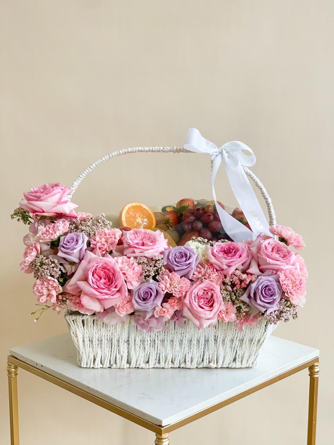 A rectanglular woven basket filled with an arrangement of pink and purple roses, finished with a large white bow on the handle. This sweet floral fruit basket also contains an assortment of fruits, including grapes and orange slices. It is placed on a small, square, gold-leg table.
