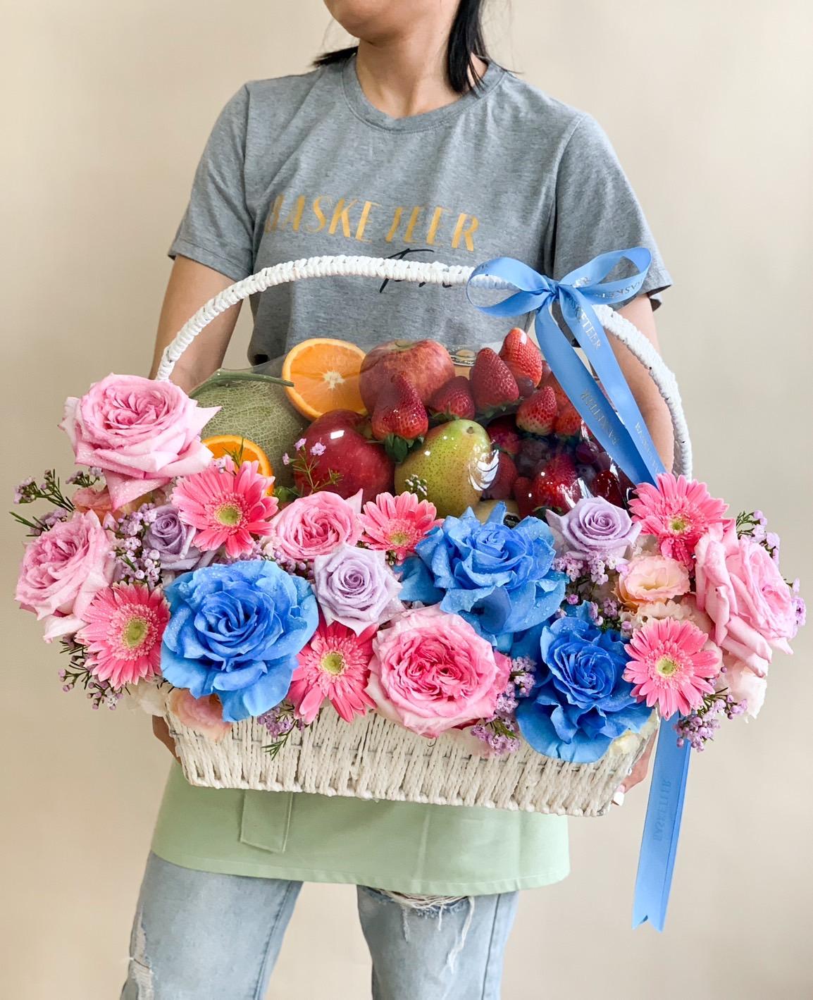 A person is holding a large white basket filled with an assortment of vibrant fresh fruits and surrounded by an array of colorful flowers, including pink, blue, and peach roses. This sweet floral fruit basket is adorned with a blue ribbon, adding an extra touch of elegance.
