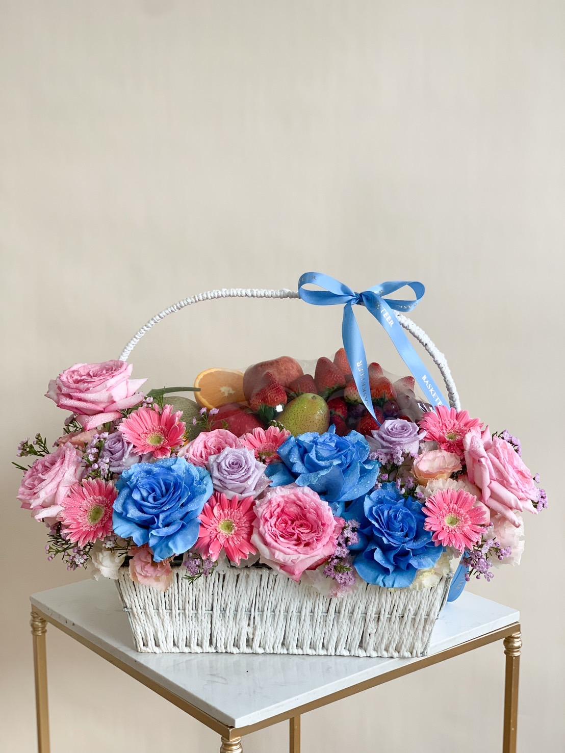 A Sweet Floral Fruit Basket, filled with a variety of fresh fruits like apples and oranges, is adorned with vibrant pink and blue roses and pink gerbera daisies. Topped with a blue ribbon, the white wicker basket rests elegantly on a small white table with golden legs.