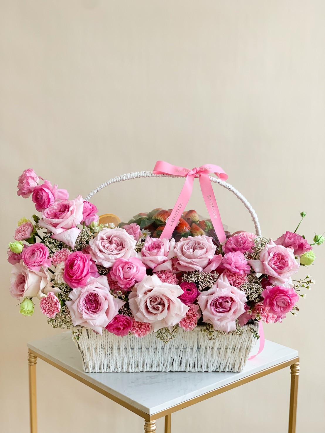 A sweet floral arrangement of pink roses and other flowers, accented with light pink and green blooms, is displayed in a white wicker basket on a small table. The charming basket, resembling a fruit basket, is adorned with a pink ribbon tied into a bow on the handle.