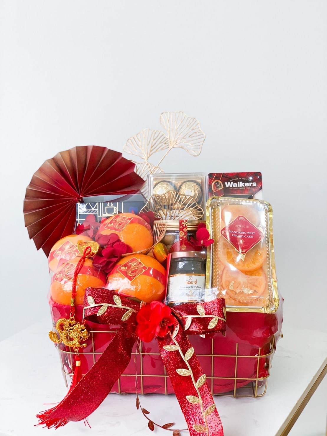 A festive red gift basket filled with oranges, a bottle of wine, Walkers shortbread cookies, and assorted chocolates. The basket is decorated with a red bow, golden fan, and intricate golden ornaments, set against a white background.