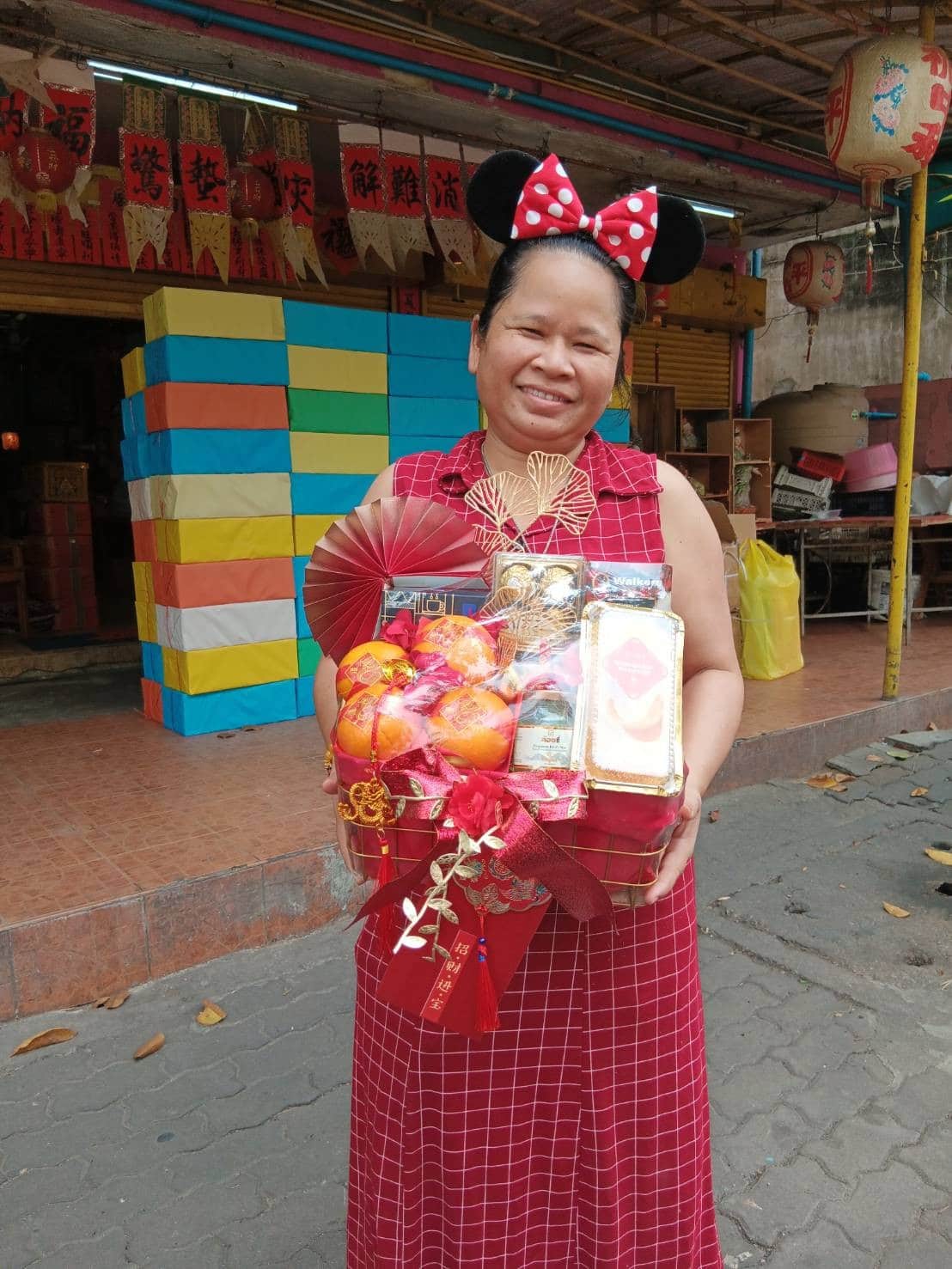 A woman wearing a red plaid dress and Minnie Mouse ears stands outdoors holding a festive gift basket containing fruit, drinks, and snacks. She is smiling and stands in front of a colorful, striped structure and a store with hanging decorations.