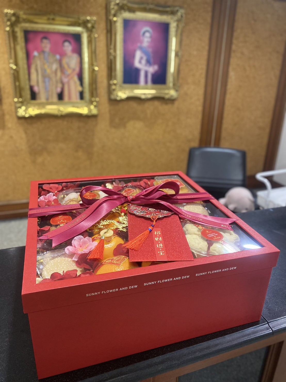 A red gift box adorned with a red ribbon and bow is displayed on a dark counter. In the box, various decorative items can be seen. In the background, two framed portraits with gold frames hang on a wooden wall beside a chair.