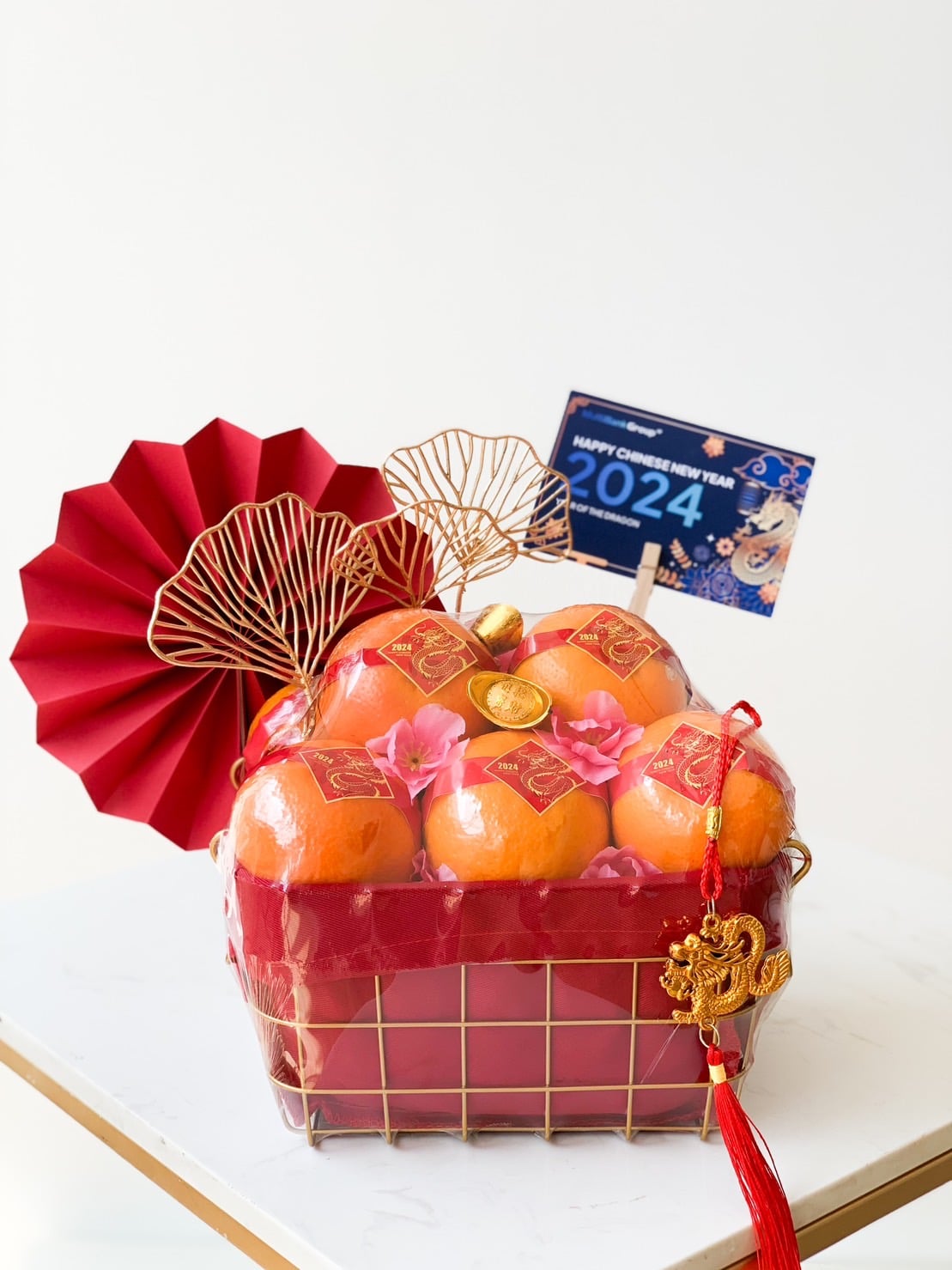 A festive gift basket filled with oranges, adorned with red envelopes and golden decorations. The basket is accented with a red paper fan and a sign saying 
