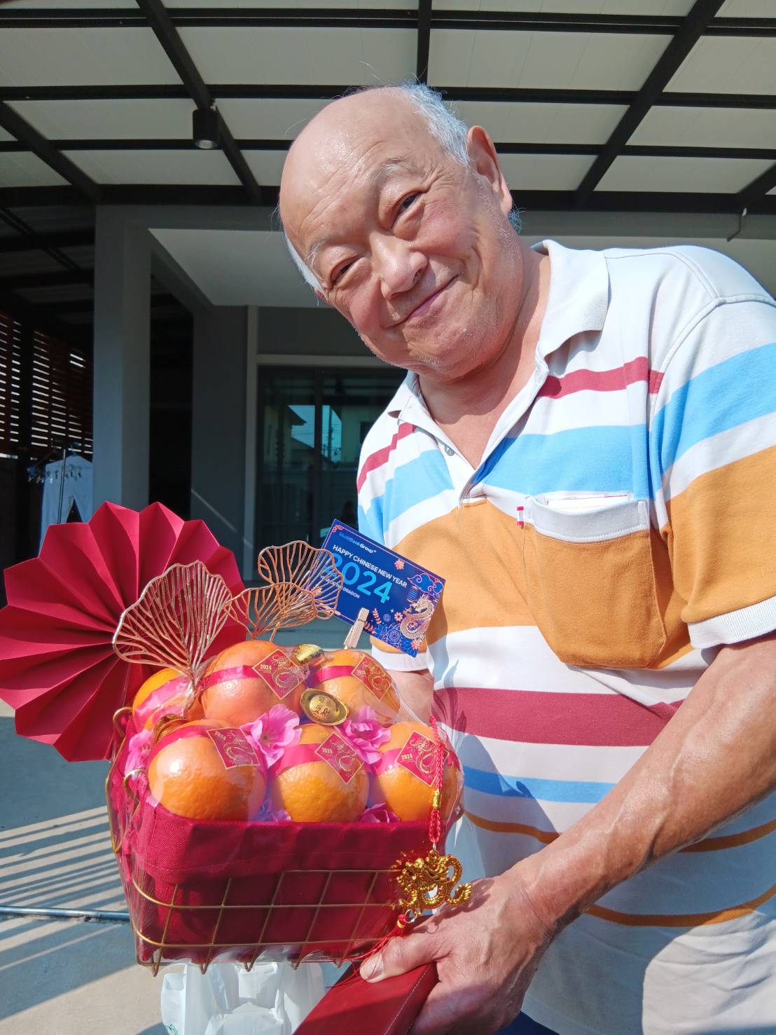 An elderly man with a bald head and a striped polo shirt is smiling and holding a decorated basket filled with oranges, red ribbons, and small ornaments. He also holds a 