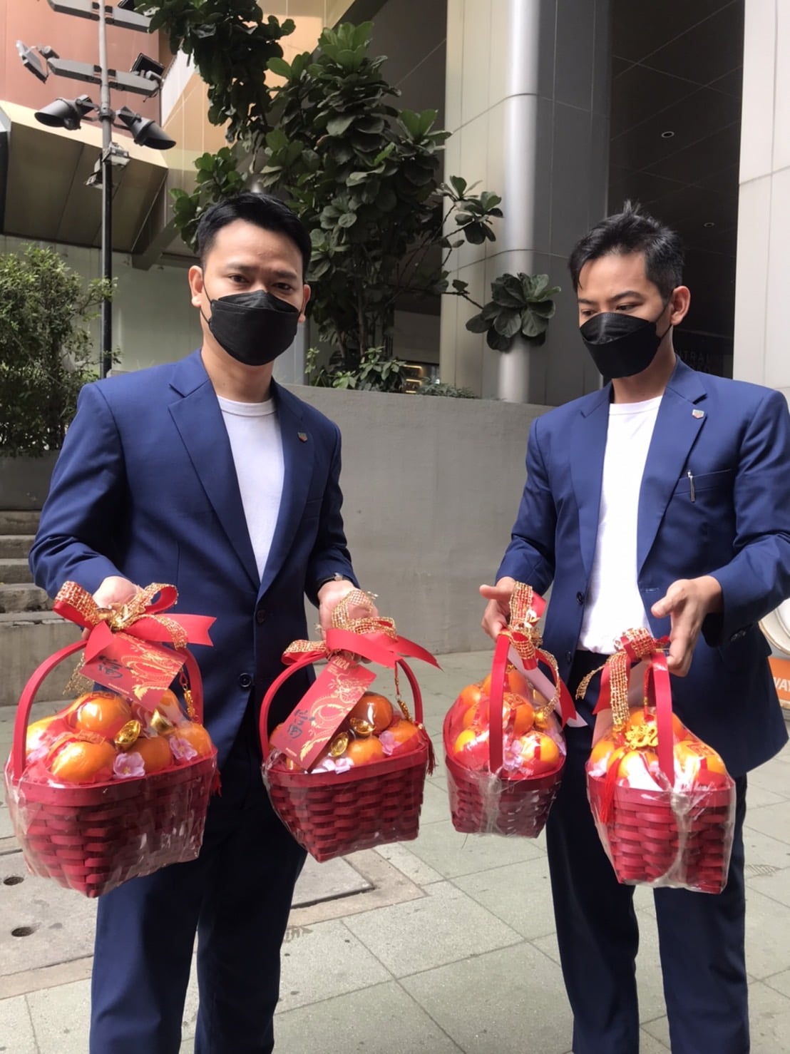 Two people in blue suits and white shirts, wearing black face masks, each holding two red woven baskets filled with fruits, likely oranges, adorned with decorative red ribbons. They are standing outdoors by a modern building and greenery.