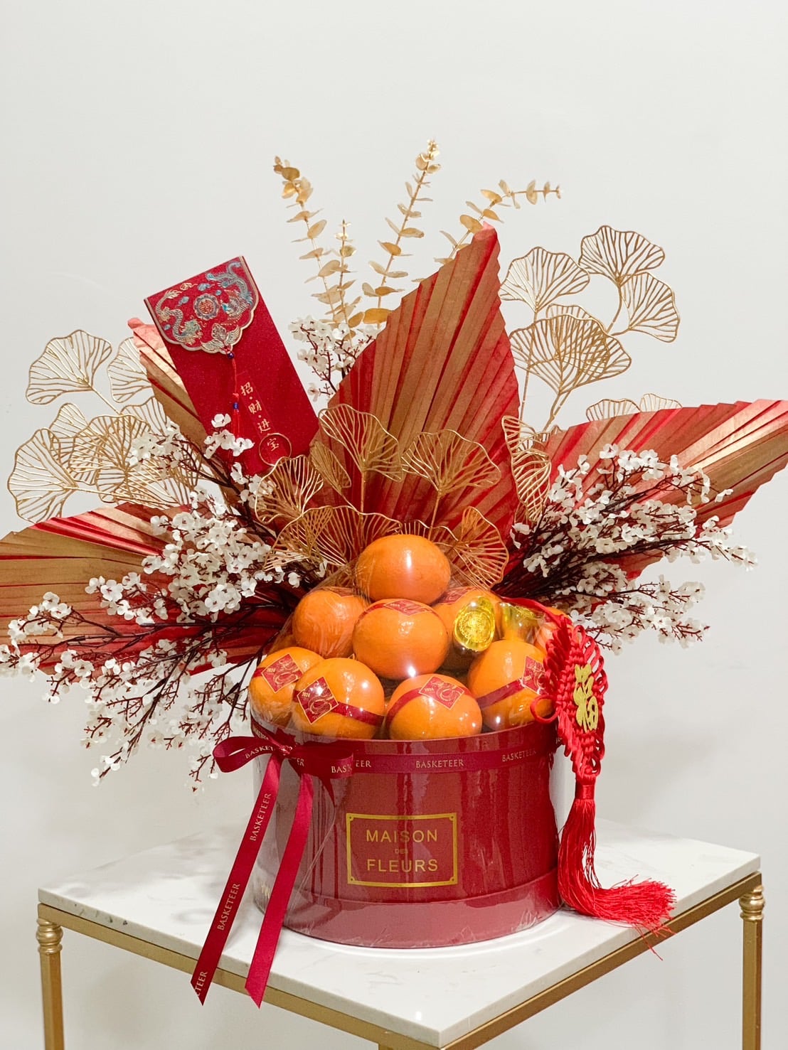A festive red arrangement featuring gold and red decorative leaves, sprigs of baby’s breath, and fresh oranges in a cylindrical container with 