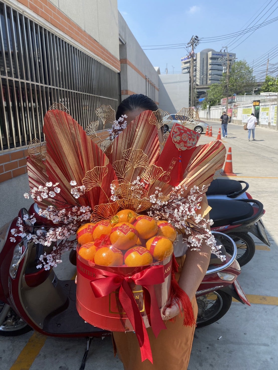 A person holding an elaborate bouquet decorated with red and gold fan-like structures, flowers, and a large arrangement of oranges. The background features a street scene with a parked scooter, buildings, and a few pedestrians.