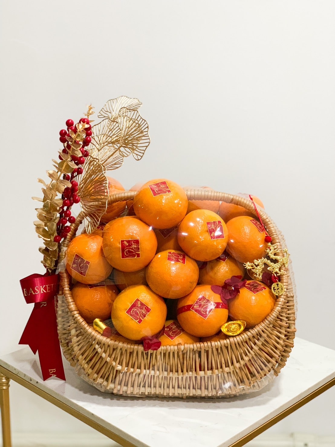 A wicker basket filled with celebratory mandarin oranges adorned with red stickers is displayed on a table. The basket is decorated with red ribbons, golden ornament leaves, red berries, and gold ingots, giving it an elegant and festive appearance.