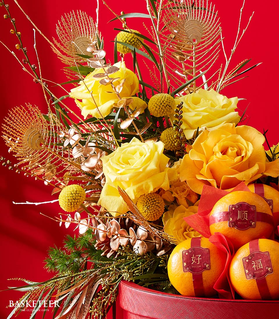 A luxurious floral masterpiece by Basketeer takes center stage, featuring radiant yellow roses and opulent golden accents, elegantly paired with spherical yellow blooms. Nestled within this vibrant arrangement are three meticulously adorned oranges, each inscribed with intricate Chinese characters that enhance the festive spirit. Set against a rich and bold red backdrop, this composition exudes an inviting sense of celebration and grandeur. The elegant presentation is further enhanced by a ribbon bearing the distinguished Basketeer logo, promising an exquisite touch to any occasion.