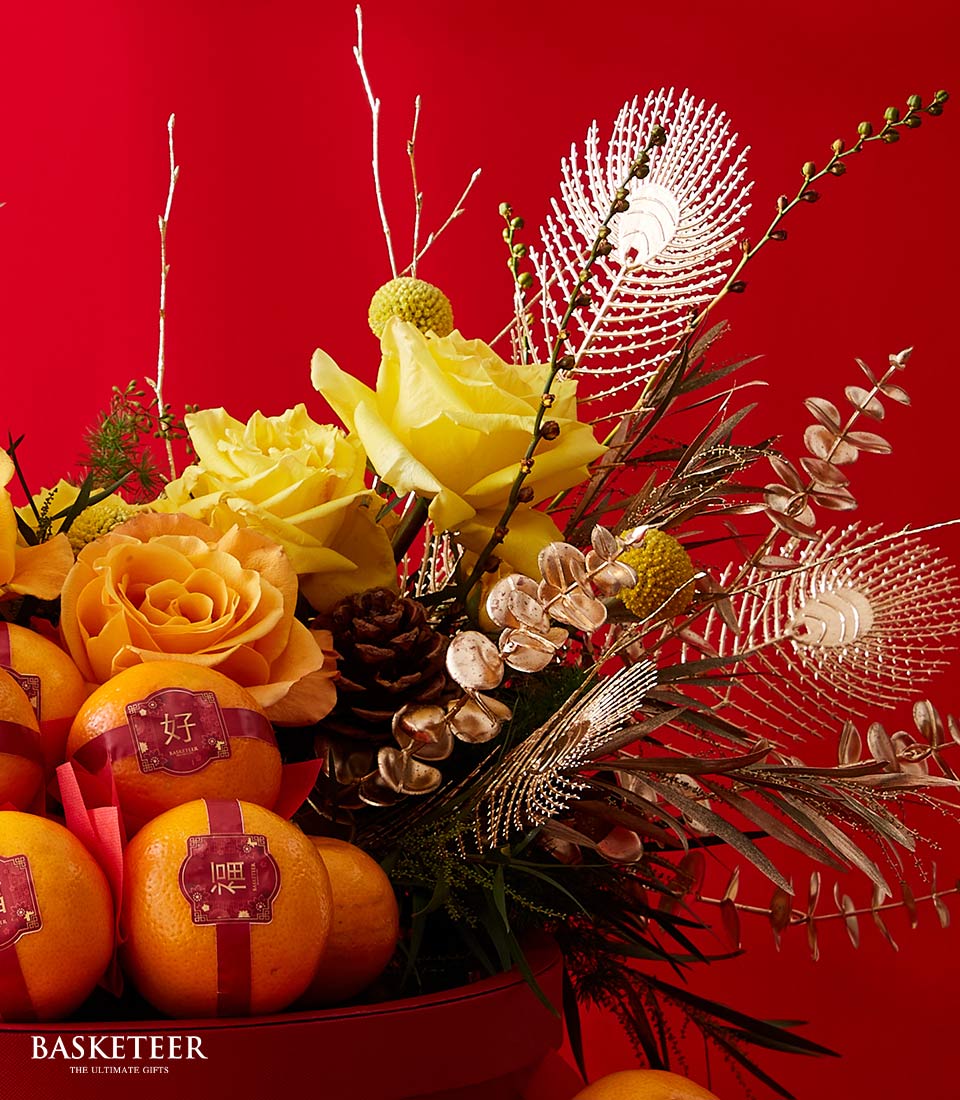 A luxurious floral arrangement graces a rich, red backdrop, the perfect embodiment of elegance and festivity. This exquisite display features radiant yellow roses paired with a delightful medley of delicate green and white blooms. Interspersed are charming pinecones and shimmering metallic accents that catch the light just so. At the base, fresh oranges adorned with red labels add a touch of natural vibrancy. The refined 
