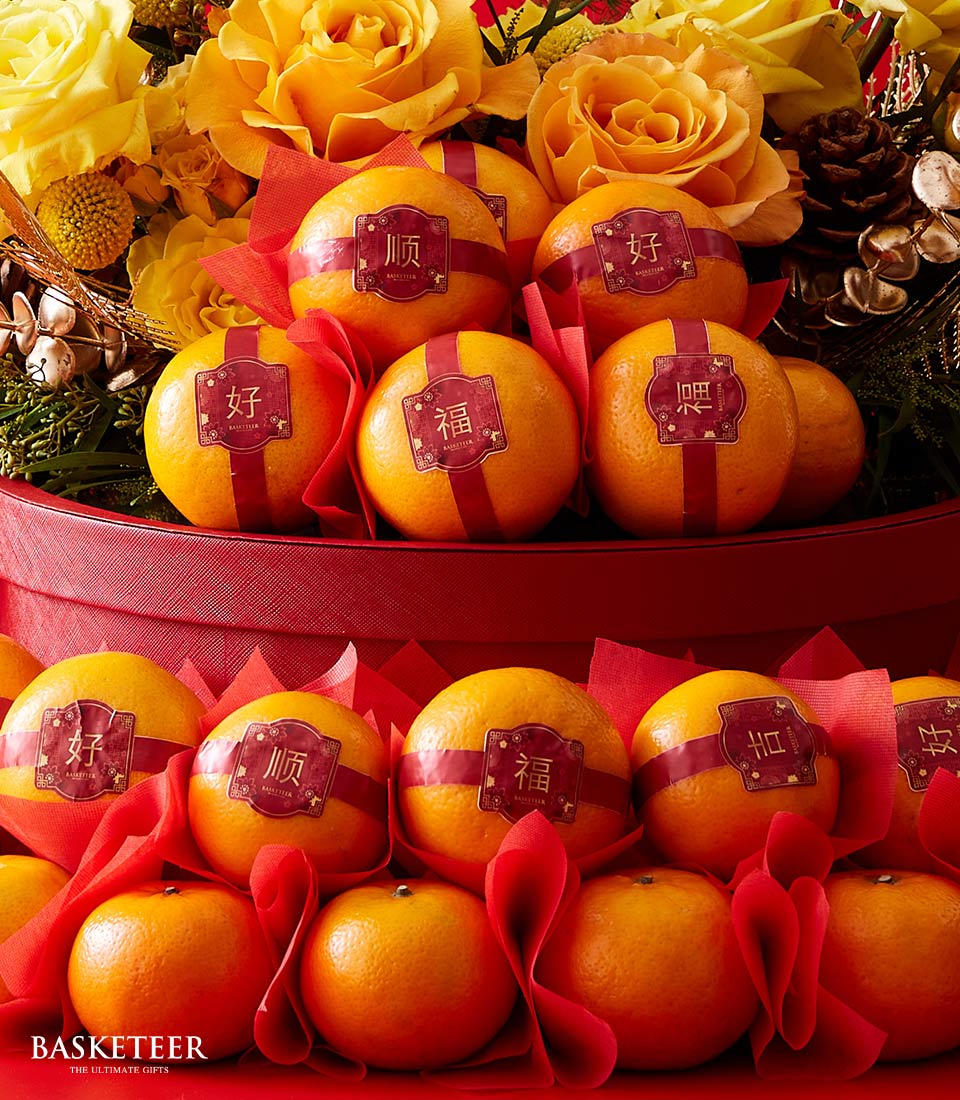 A lavish display of sumptuous oranges, each elegantly adorned with red ribbons and Chinese characters, meticulously arranged in two luxurious tiers lined with exquisite red fabric. Surrounding the arrangement are vibrant yellow roses, charming pinecones, and lush leafy greens, all perfectly curated to evoke an opulent and festive ambiance. A sophisticated touch bearing the Basketeer logo enhances the scene's refinement.