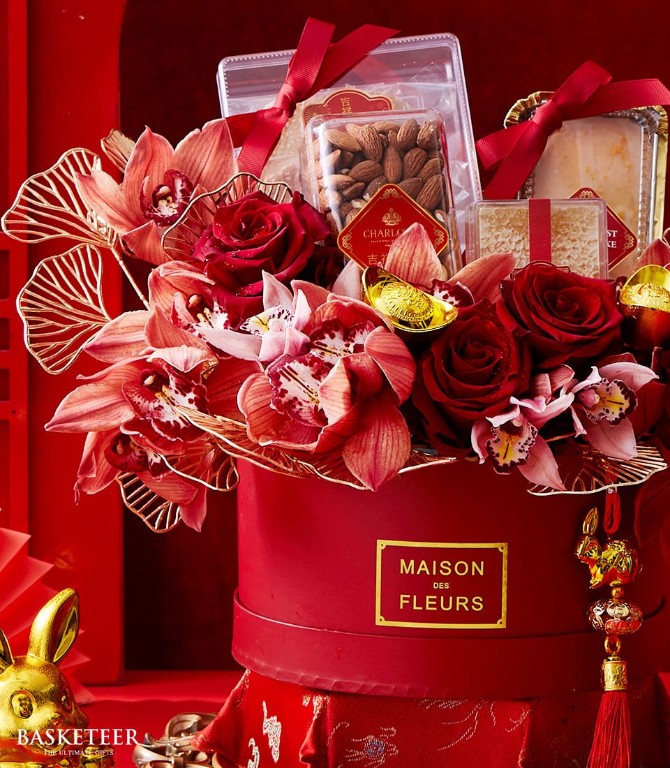 A lavish red gift basket, exquisitely curated by Basketeer, overflowing with an array of delectable treats. Nestled within are premium packaged almonds, sumptuous cheese, and decadent chocolate elegantly wrapped in gold foil. The scene is further enriched by an enchanting assortment of vibrant red and pink flowers. Accentuated with opulent ribbons and adorned with the distinguished label 