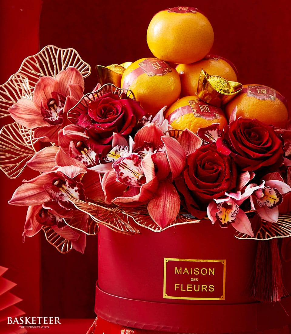 A lavish ensemble showcasing resplendent red roses, graceful pink orchids, and radiant golden-hued oranges, all artistically arranged within a sophisticated red circular box branded 