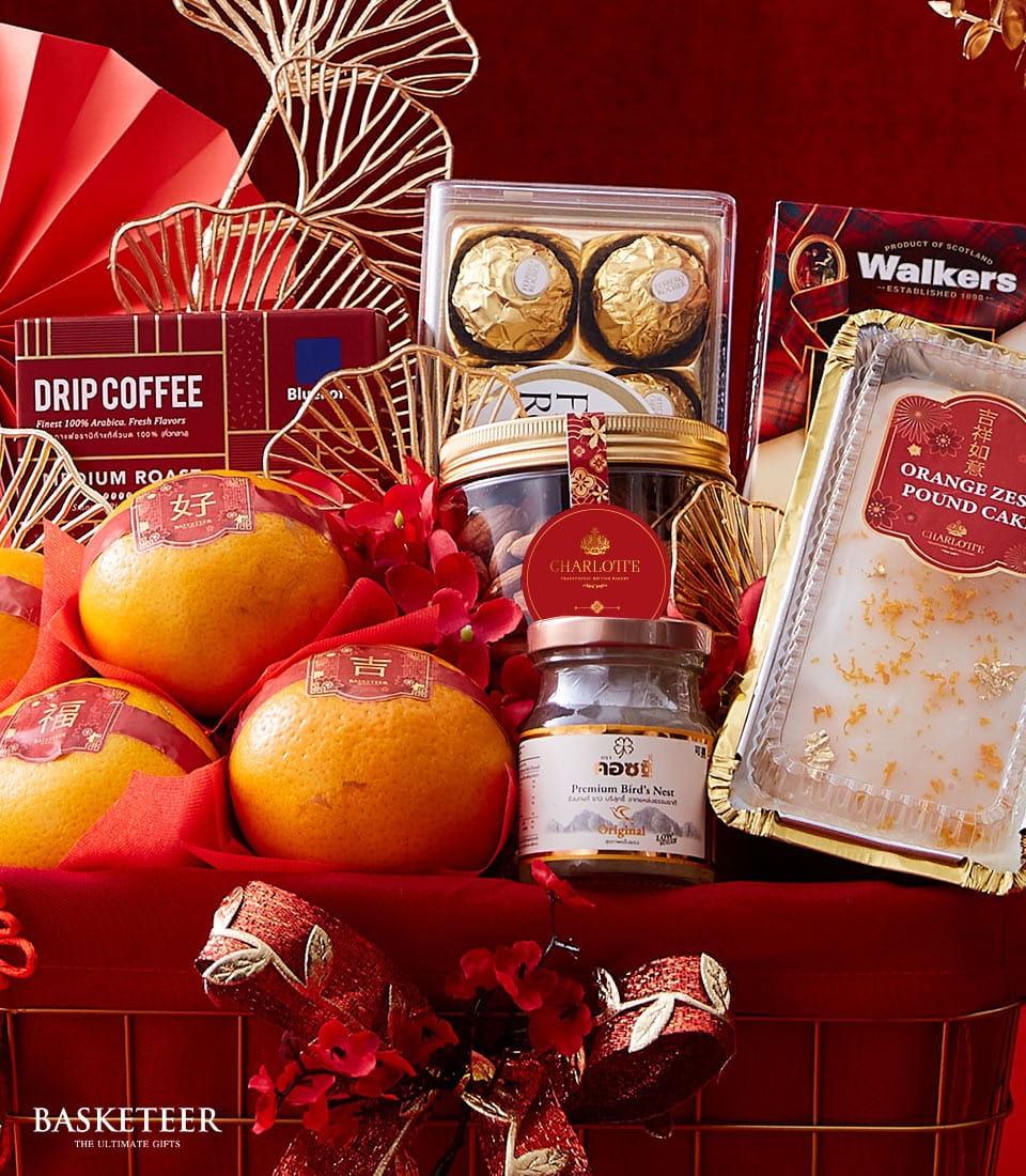 A lavishly curated gift basket from Basketeer, graced with vibrant oranges, gourmet drip coffee packets, decadent Ferrero Rocher chocolates, buttery shortbread cookies, a fragrant orange zest pound cake, and a jar of premium mixed nuts. The basket exudes elegance with its red and gold accents, delicate flowers, and an exquisite ribbon. The distinguished BASKETEER logo is prominently displayed at the bottom, epitomizing luxury and indulgence.