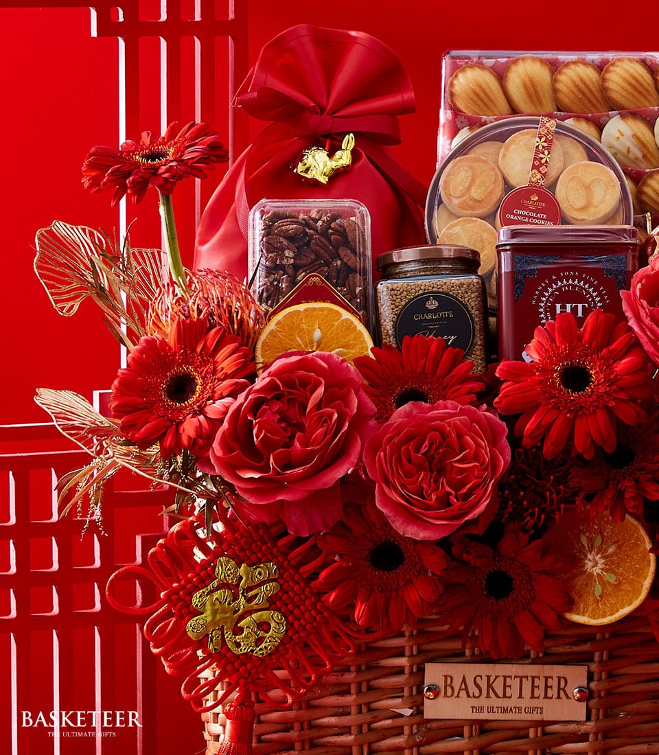 A lavishly curated gift basket by Basketeer, brimming with exquisite red flowers and zesty dried orange slices. This opulent arrangement includes an assortment of delectable packaged treats such as gourmet nuts, artisanal cookies, and premium tea. The basket is elegantly adorned with rich red and gold accents, topped with a luxurious red fabric pouch. The distinguished Basketeer logo is prominently displayed, completing the scene with a touch of refined sophistication.