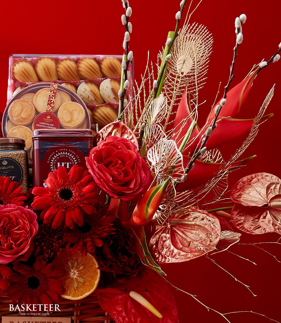An opulent Basketeer gift basket set against a sumptuous red backdrop features an enchanting arrangement of festive flowers, including lush red roses and vibrant gerberas, interwoven with luxurious dried elements. This exquisite basket is brimming with decadent premium biscuits and an assortment of gourmet snacks, elegantly garnished with slices of zesty orange. The prestigious Basketeer label graces the bottom left corner, adding a touch of sophistication to this lavish display.