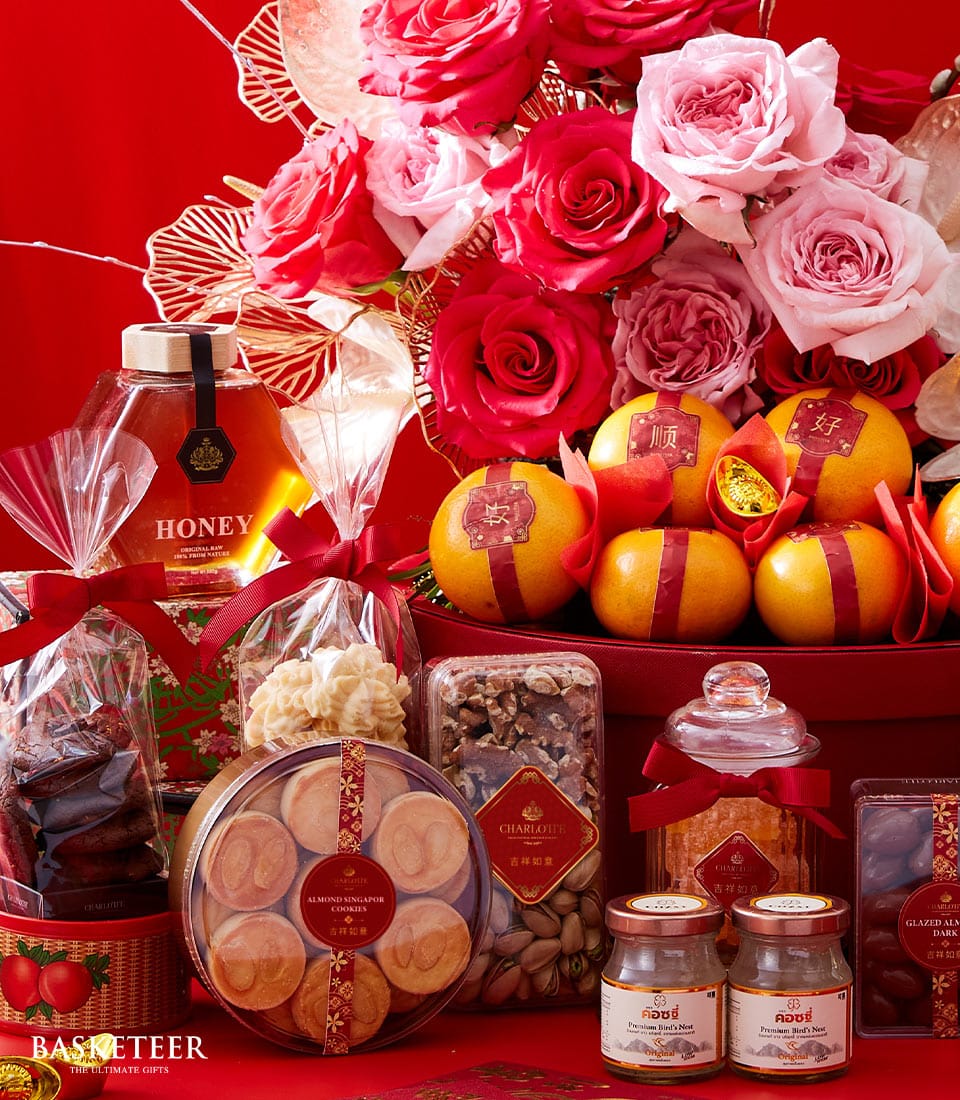 A luxurious Basketeer gift basket brimming with succulent oranges, artisanal honey jars, an assortment of gourmet nuts, exquisite cookies, and delectable snacks. Nestled within elegant packaging adorned with the Basketeer logo, the scene is further elevated by a stunning arrangement of pink and red roses. Set against a vibrant red backdrop, the display exudes festive opulence and celebratory sophistication.