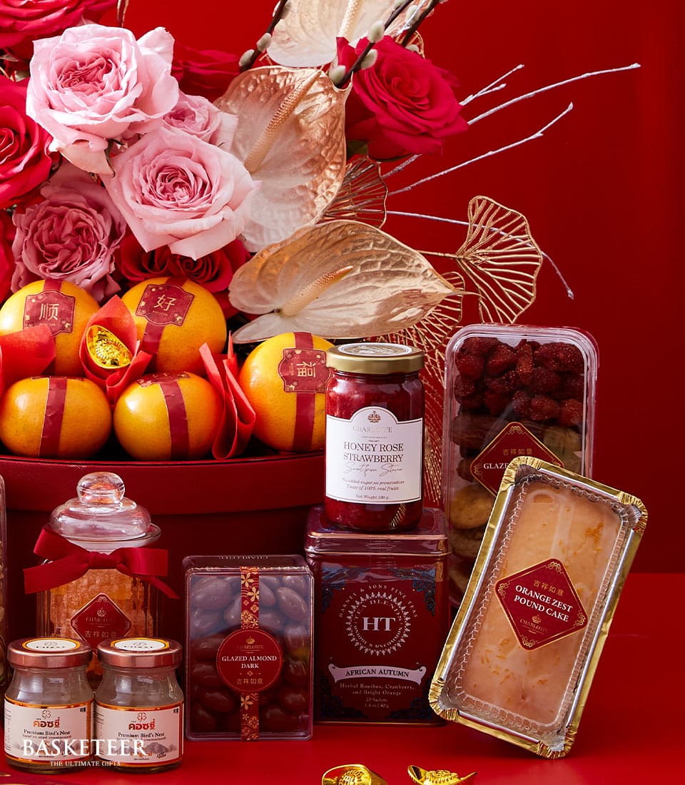 An opulent Basketeer gift basket set against an elegant red backdrop. The meticulously arranged basket boasts a bouquet of delicate pink roses, alongside a selection of crisp yellow and red apples. Nestled within are gourmet delights such as a jar of honey rose strawberry preserves, a package of sumptuous glazed Medjoul dates, and an assortment of other exquisite preserves and baked goods. A ribbon featuring the Basketeer logo elegantly adorns the arrangement, signaling an unmatched experience in luxury gifting.