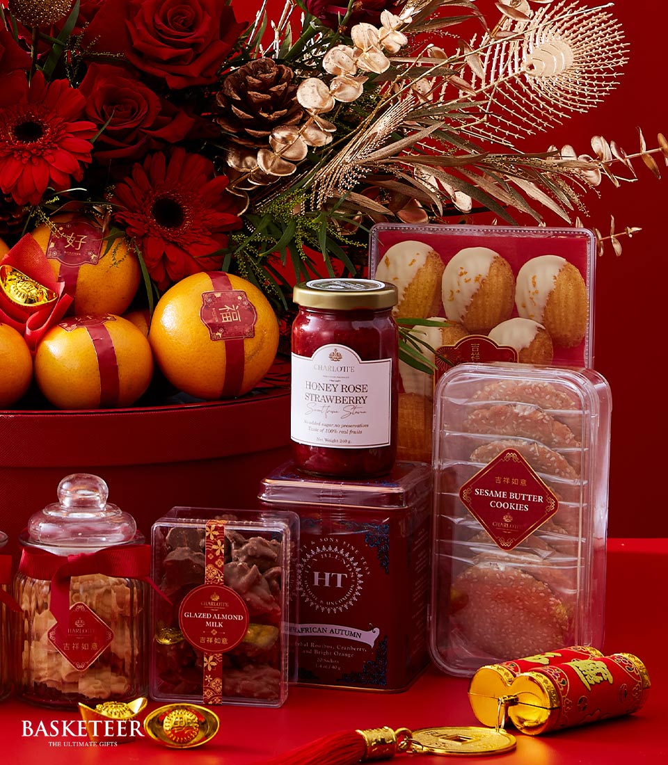 An opulent festive gift arrangement featuring a luxurious red basket adorned with the prestigious Basketeer logo, brimming with succulent oranges, a jar of exquisite honey rose strawberry preserves, delectable assorted cookies, gourmet nuts, and a gleaming small gold ingot. This elegant setup is enhanced by lavish red and gold decorative elements and delicate floral embellishments, all set against a rich red backdrop that exudes warmth and sophistication.
