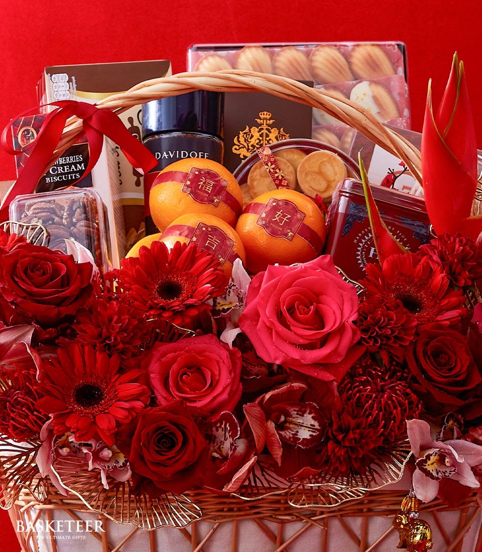 A luxurious Basketeer gift basket beautifully adorned with vibrant red flowers and premium oranges, each marked with a sophisticated red label. Nestled among the fresh fruits are elegantly boxed biscuits and an assortment of decadent chocolates, all meticulously arranged to perfection. The entire ensemble is set against a rich red backdrop, exuding an air of festive opulence. A ribbon tastefully displaying the Basketeer logo adds the final touch of refinement to this inviting scene.