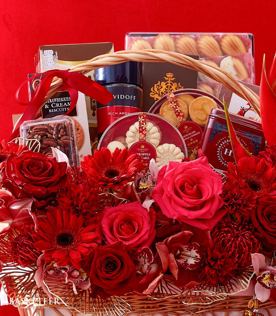 A luxurious Basketeer gift basket brimming with an exquisite assortment of gourmet treats, elegantly presented and adorned with vibrant red flowers. Inside this opulent arrangement, you'll discover beautifully packaged boxes of fine biscuits, decadent chocolates, and premium coffee. Each item is meticulously wrapped in sumptuous ribbons bearing the distinguished Basketeer logo. Set against a rich red backdrop, this lavish display is the epitome of sophistication—perfect for Valentine's Day or any romantic occasion seeking an unforgettable touch of elegance.