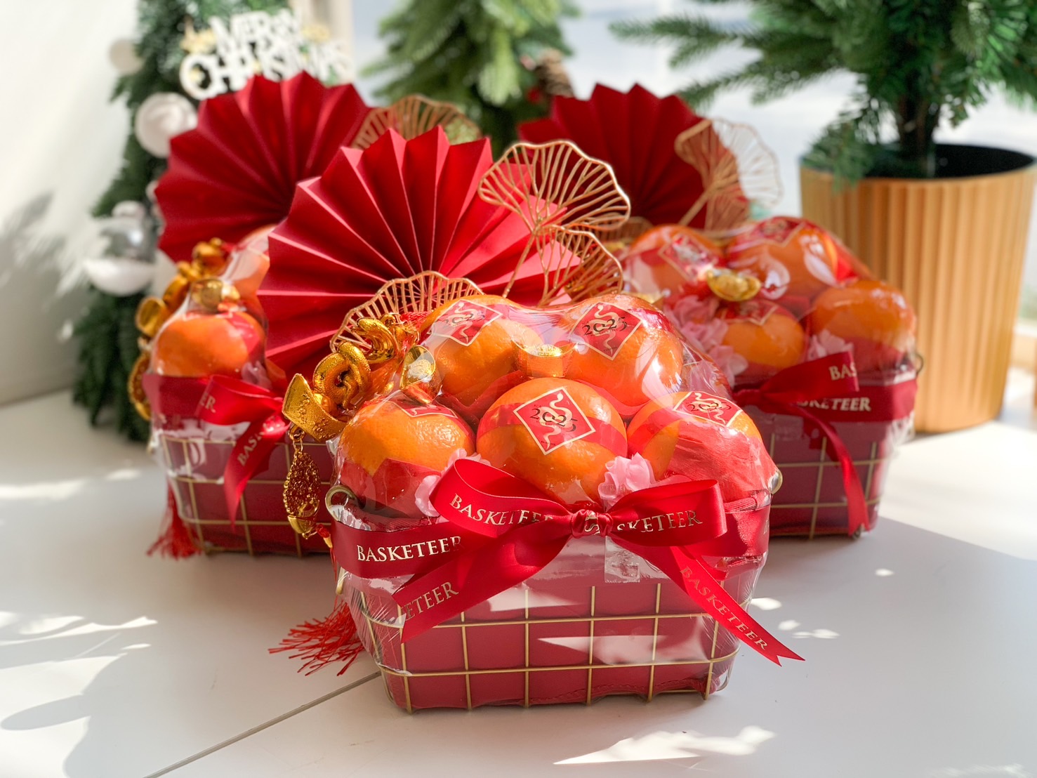 Beautifully designed Chinese New Year hampers featuring fresh oranges, gold wire baskets, red festive fans, pink blossoms, and golden charms, perfect for gifting during the celebrations.