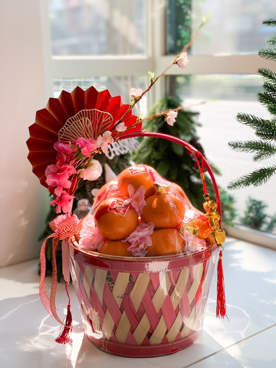 Red woven basket filled with tangerines, cherry blossoms, and a decorative fan, ideal for Chinese New Year celebrations.