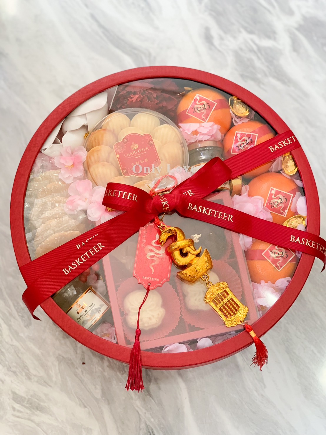 Deluxe red round Chinese New Year gift box with festive ribbons, golden charms, mandarin oranges, and assorted treats.